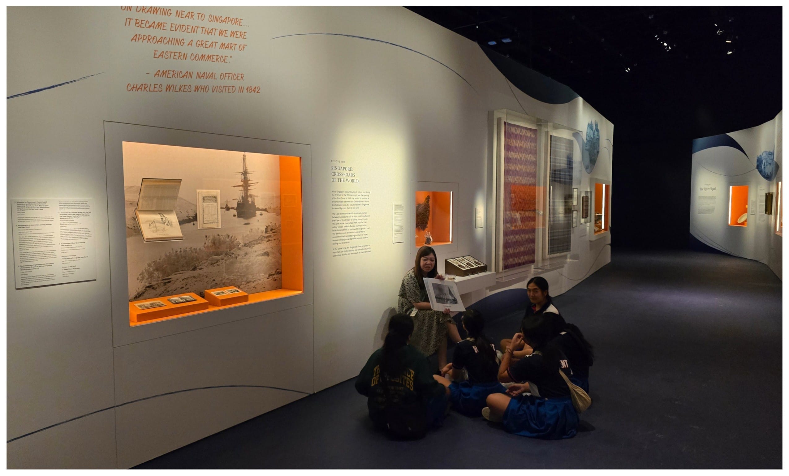 A group of students sit on the floor in a museum gallery about Singapore, while a guide holds up a photograph.