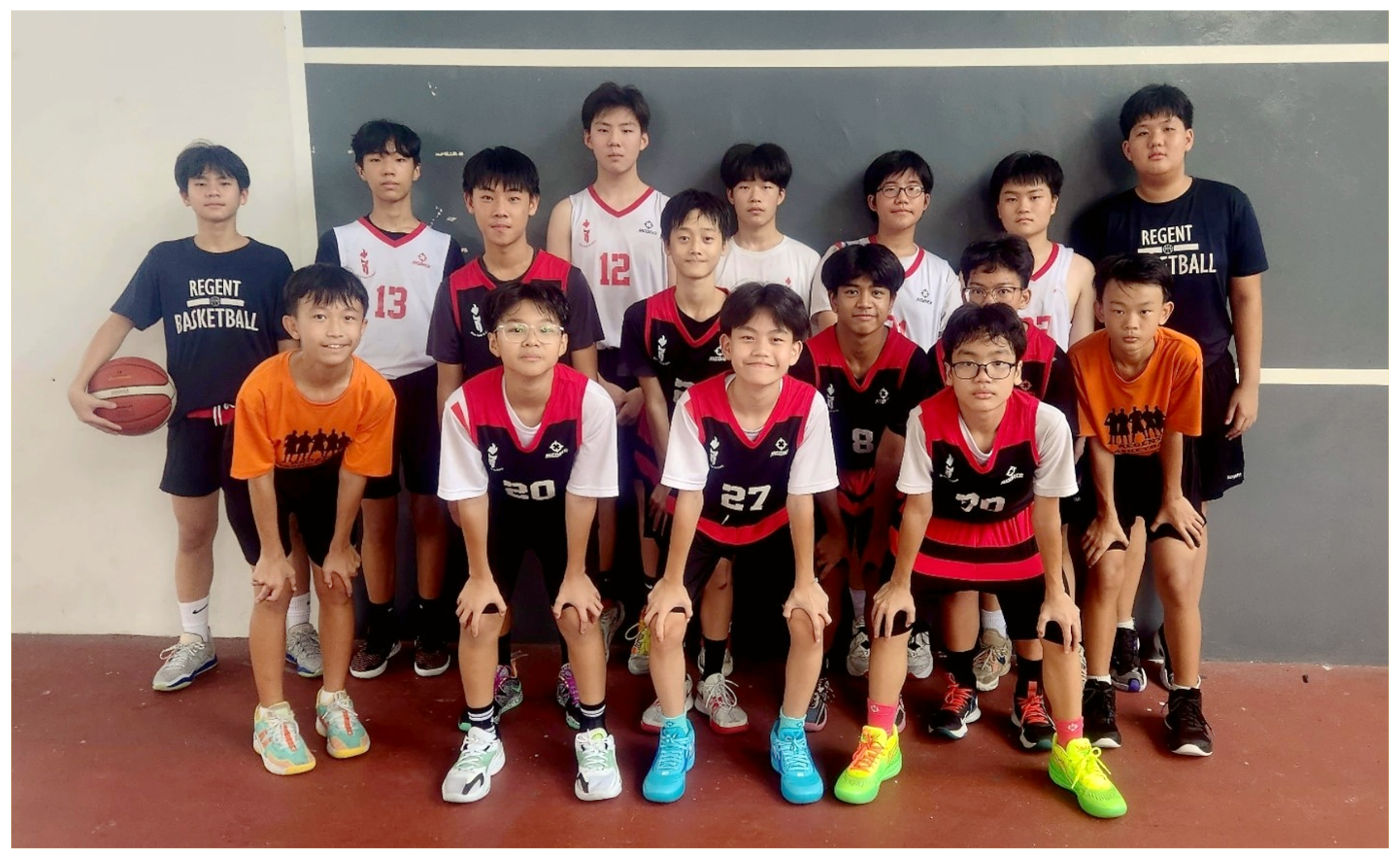 Group photo of 14 boys in basketball jerseys, some crouching, one holding a ball, on an indoor court.