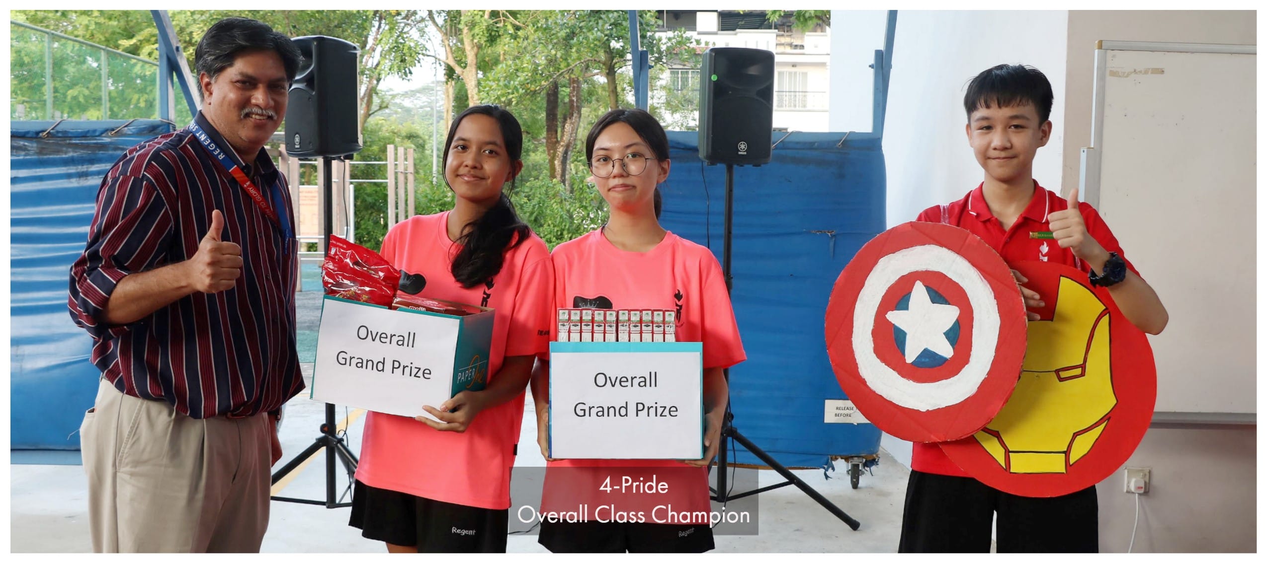 Four people pose outdoors holding Overall Grand Prize boxes and superhero shields, captioned 4-Pride Overall Class Champion.