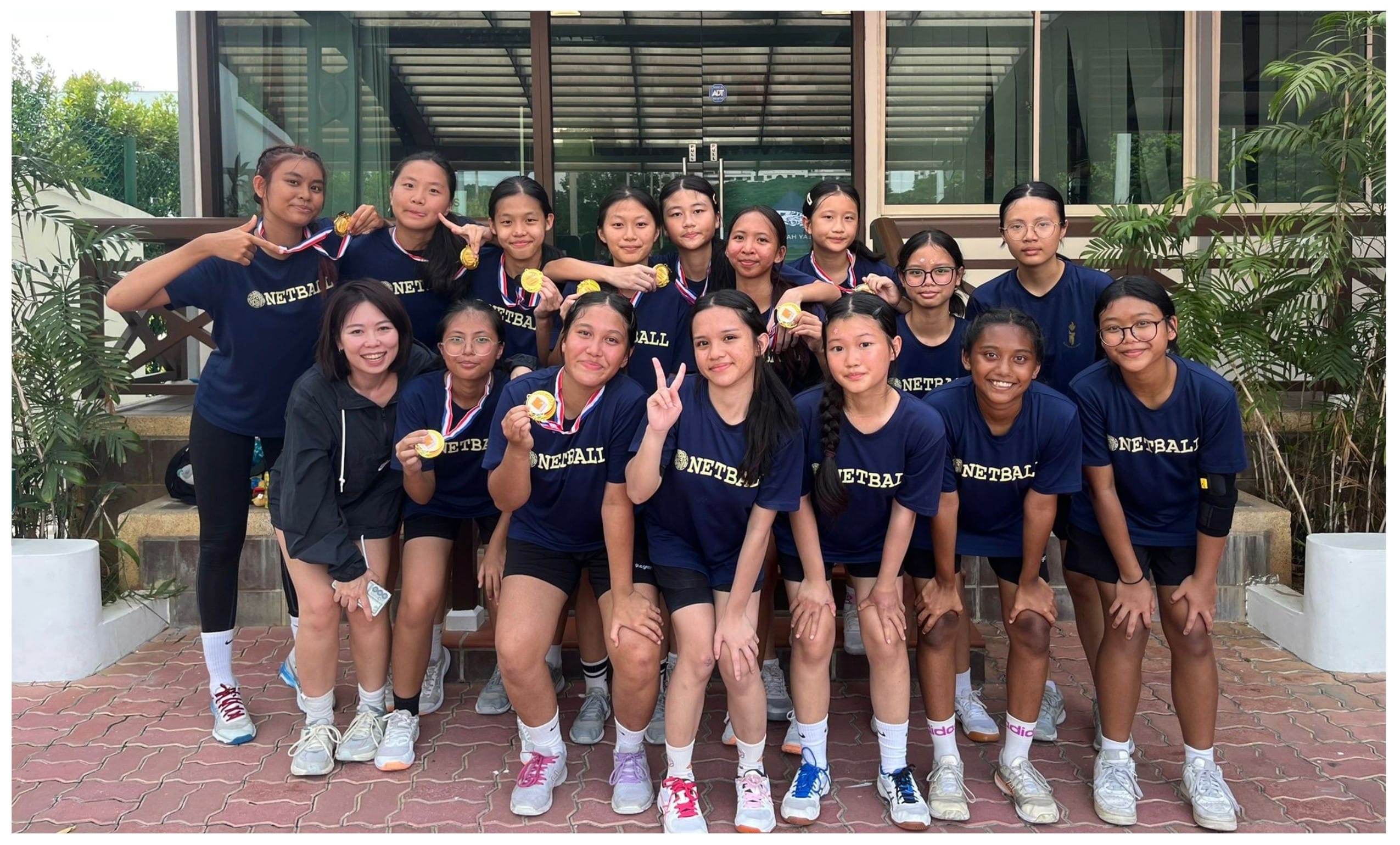A youth netball team in navy shirts poses outdoors, proudly displaying gold medals with a coach beside them.
