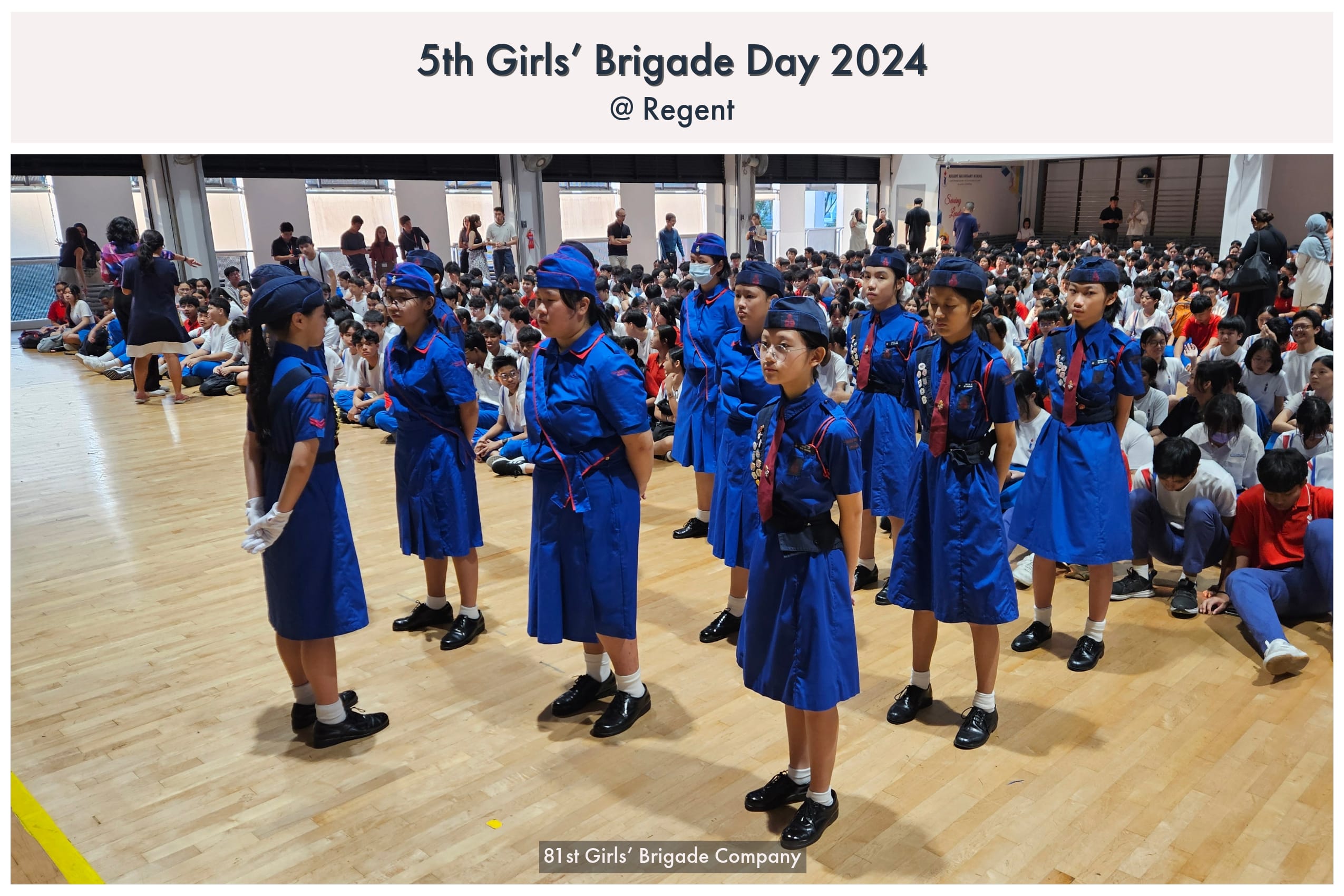 Girls in blue uniforms and caps stand in formation on a gymnasium floor at the 5th Girls' Brigade Day 2024.