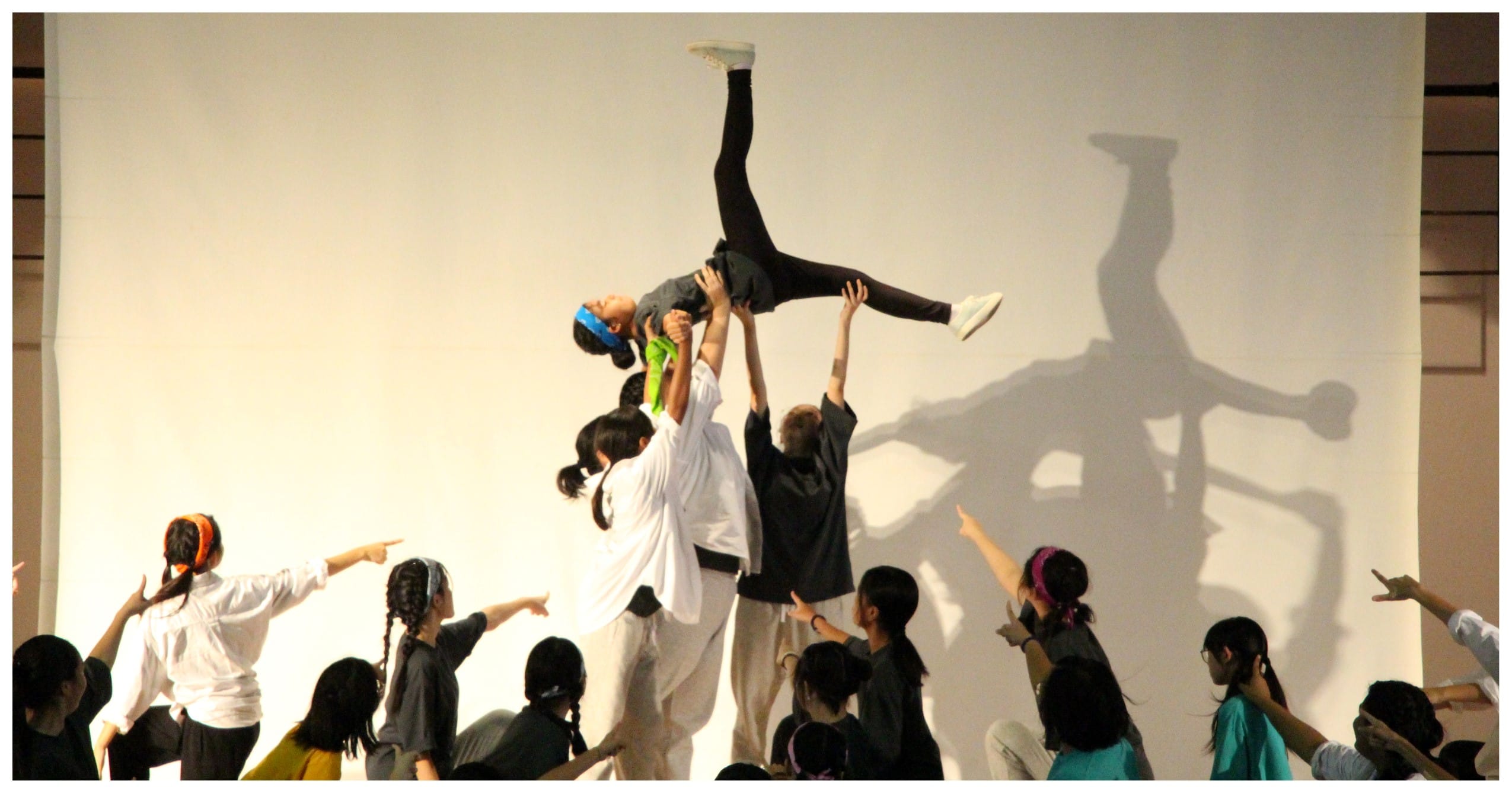 A group of young dancers lifts one performer high overhead during a stage performance, with shadows cast on a white backdrop.
