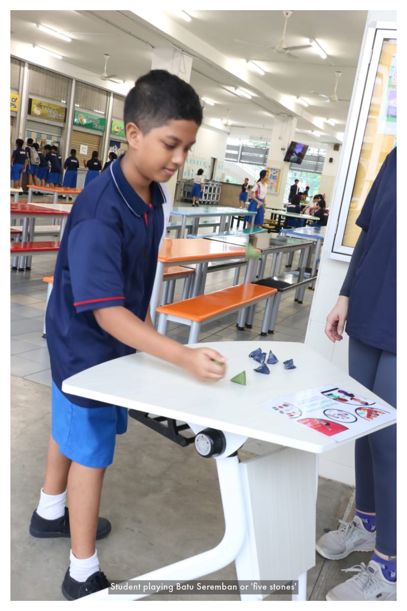 A boy in a blue school uniform plays Batu Seremban with small pyramid-shaped stones on a white table.
