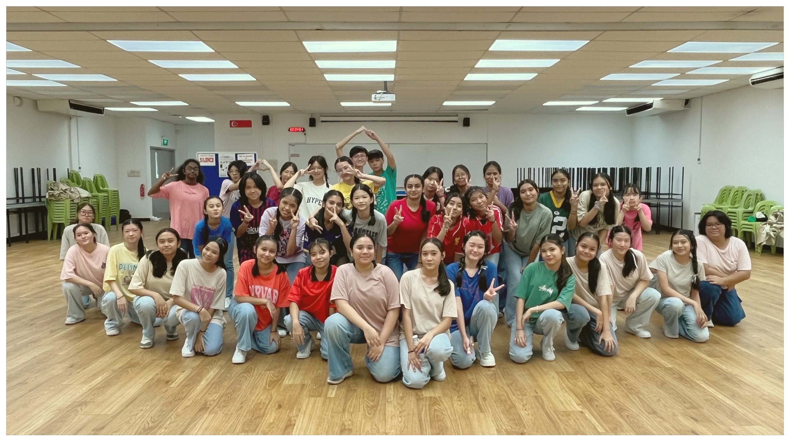 A large group of young people posing together in an indoor hall, many making peace signs and smiling.