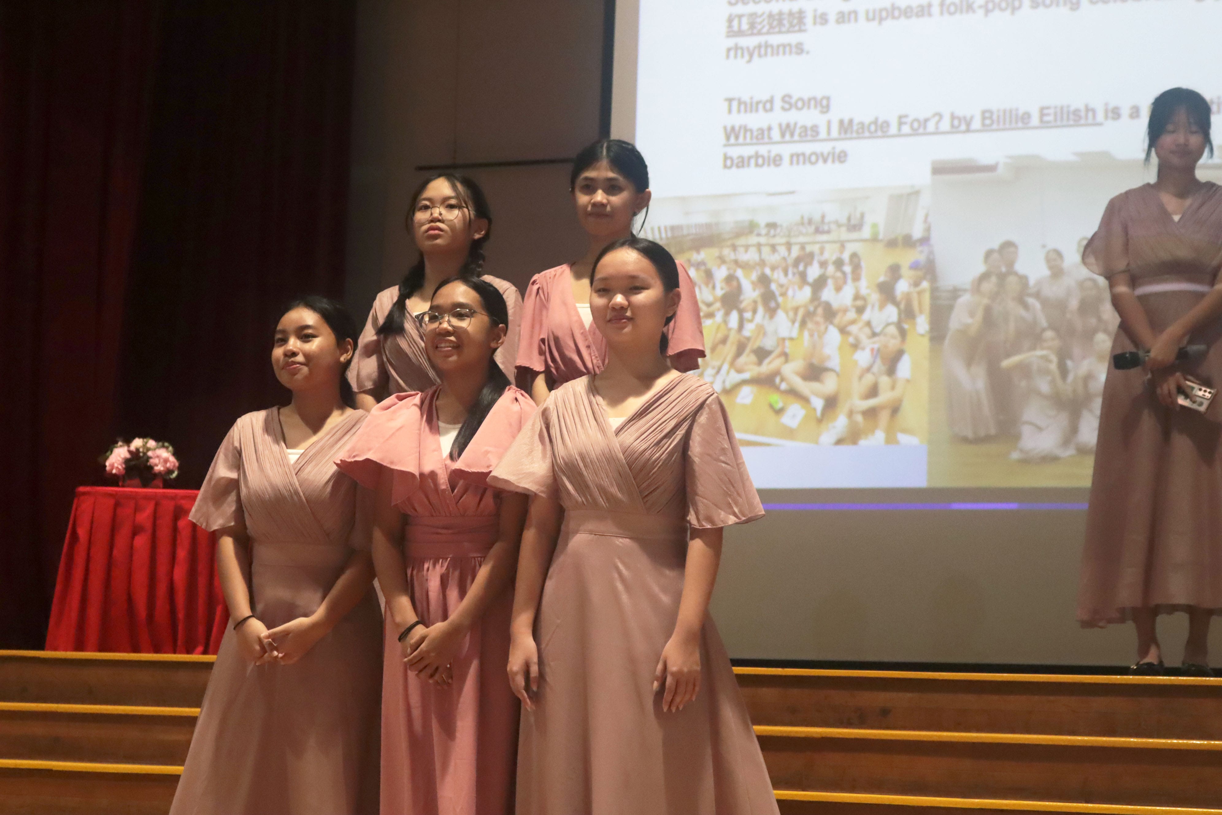 Six young women in pink and mauve dresses stand on a stage in front of a presentation screen.