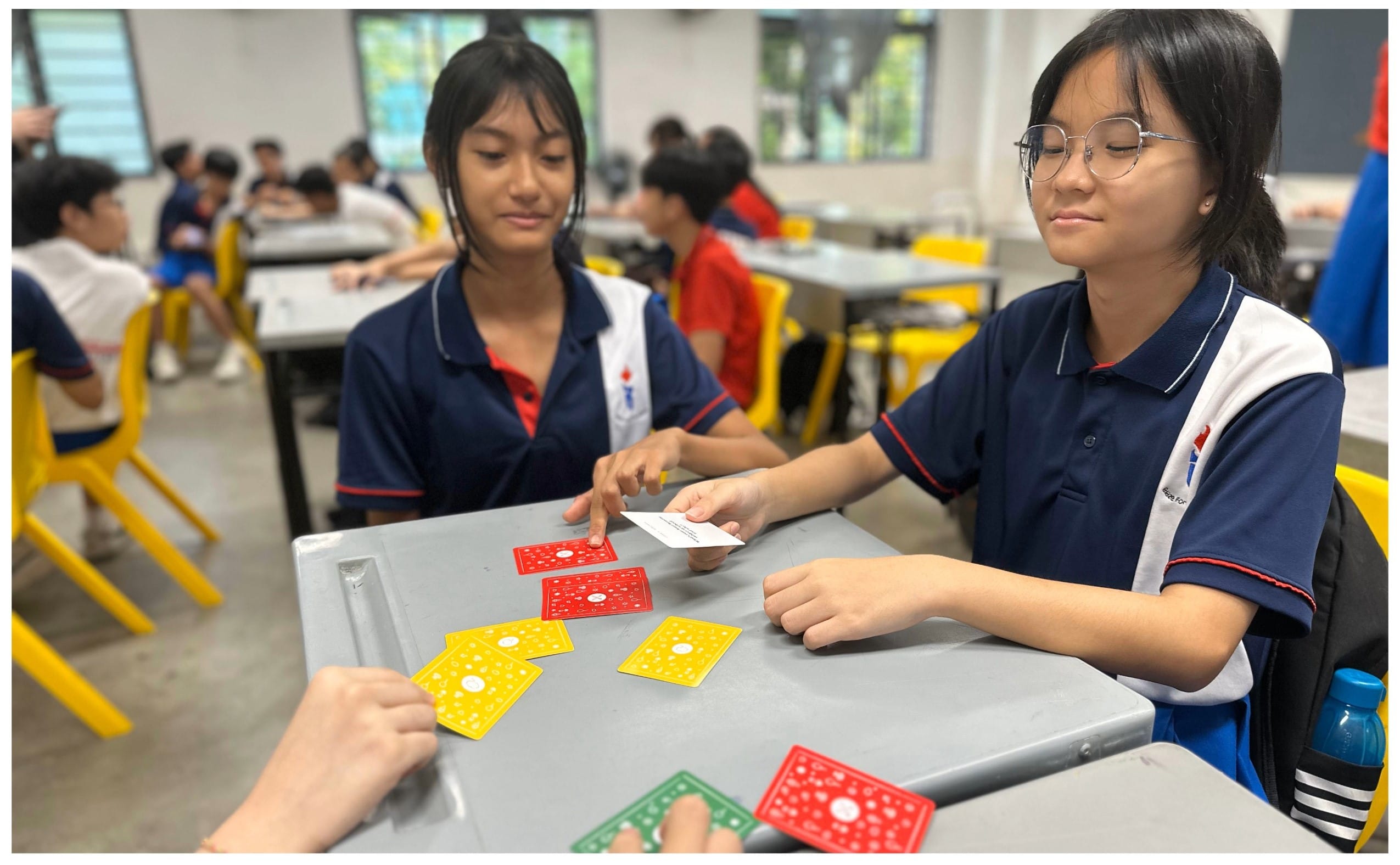 Two school girls in navy uniforms play a card game with red, yellow, and green cards at a table.