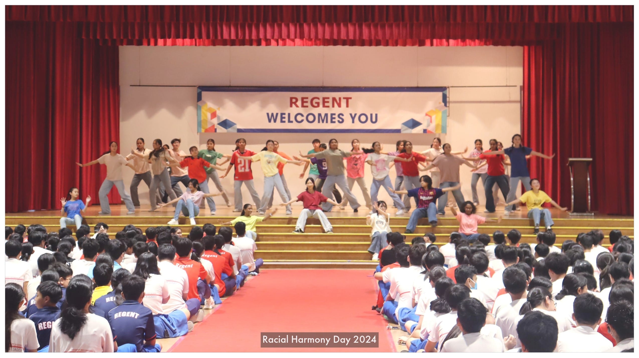 Students perform a dance on a school stage at Regent's Racial Harmony Day 2024, watched by a seated audience.