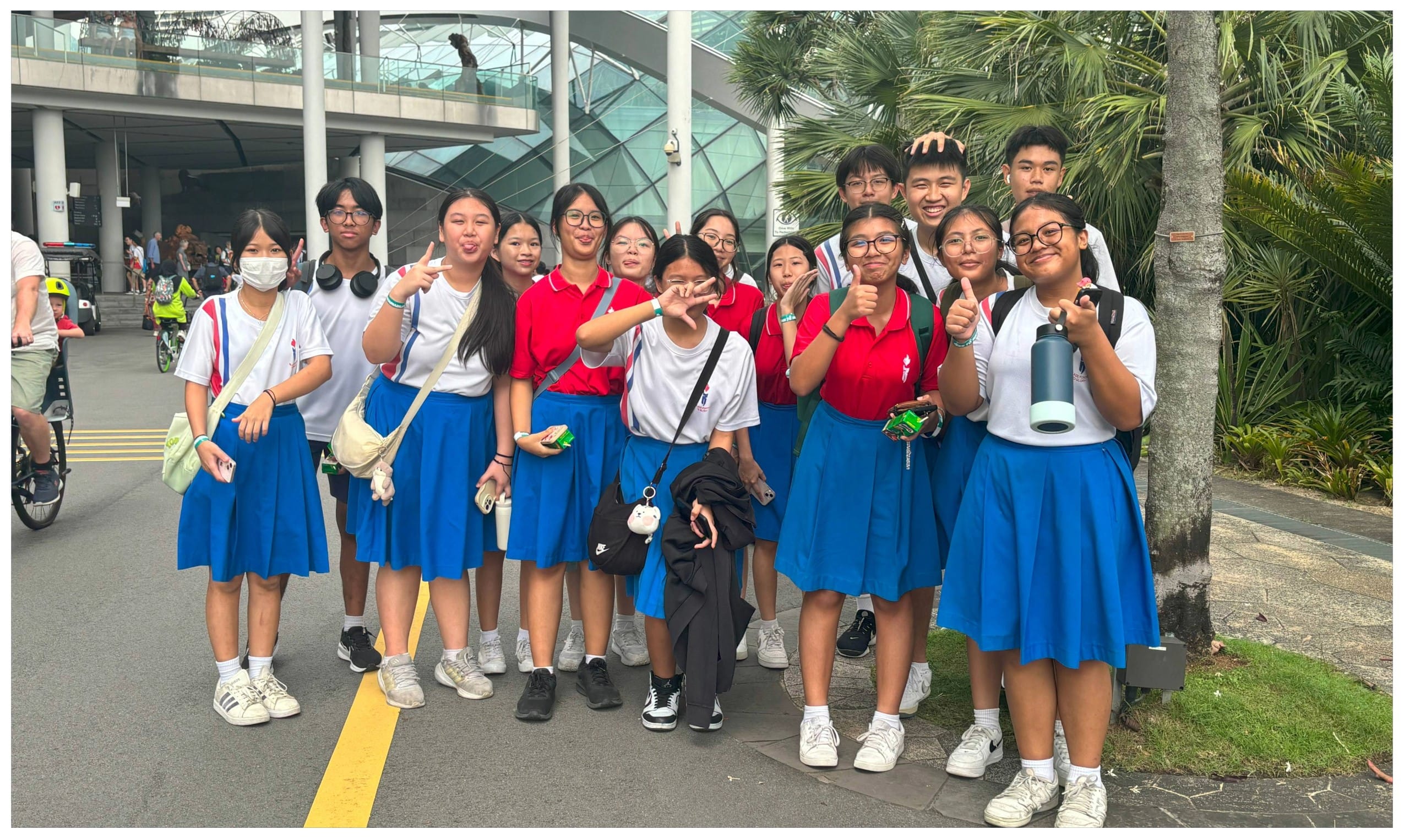 Group of students in blue skirts and red or white polo shirts posing outdoors near a modern building with palm trees.