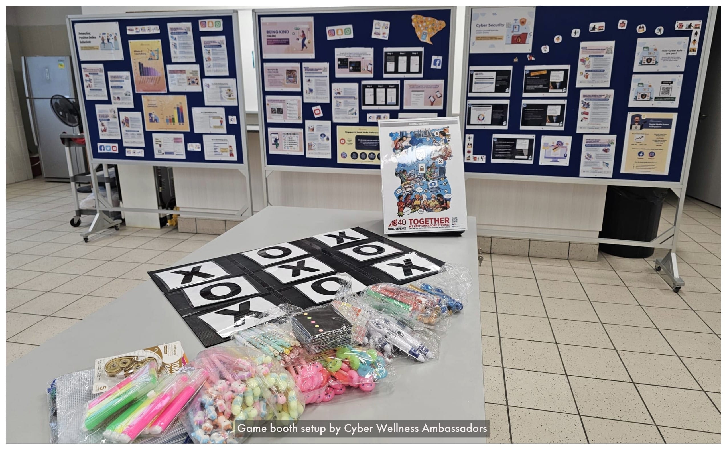A table with a tic-tac-toe mat, stationery, and prizes in front of blue notice boards with cyber wellness posters.