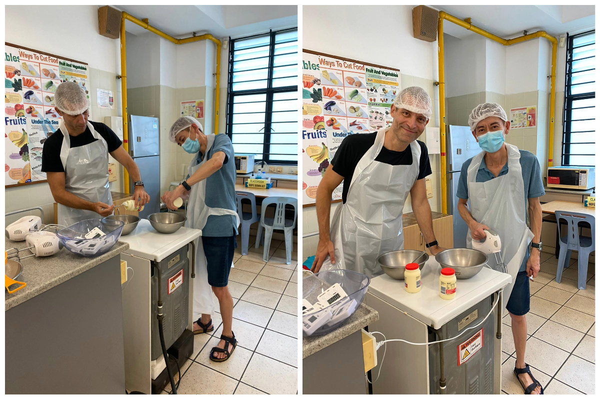 Two people wearing hairnets and aprons cook in a classroom kitchen, using mixing bowls and a hand mixer.