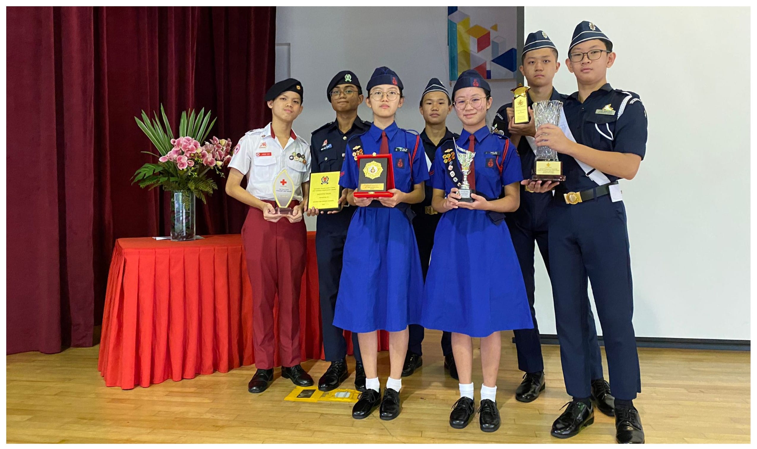 Six students in uniforms pose holding trophies and plaques on a wooden stage with a red curtain backdrop.