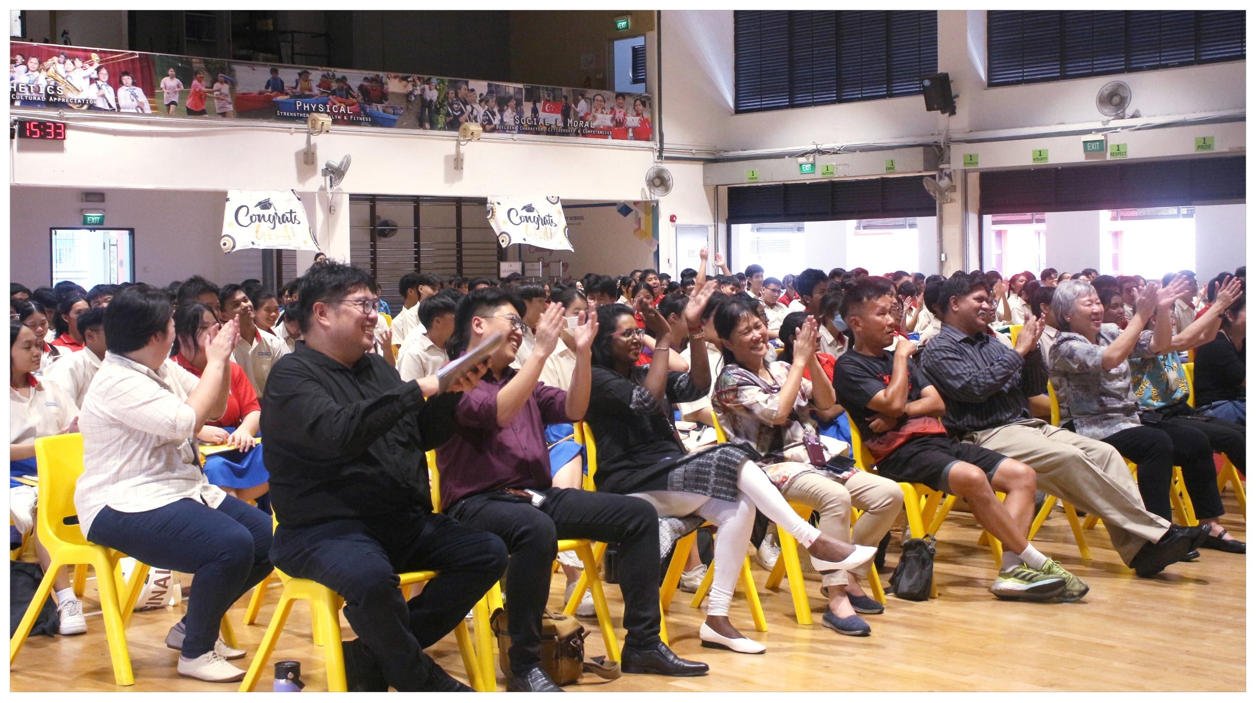 A large crowd claps and cheers on yellow chairs in a school hall with Congrats banners overhead.