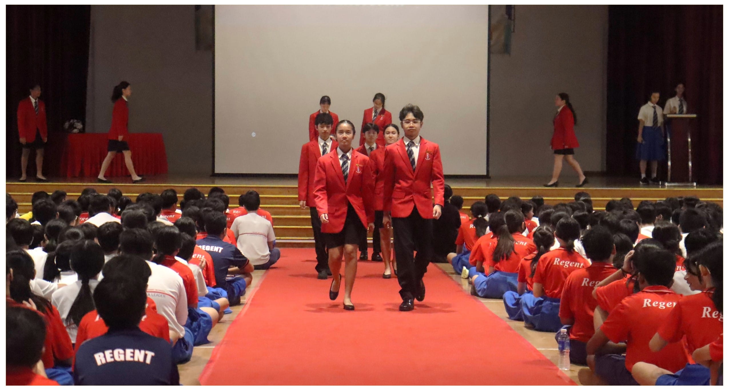 Students in red blazers walk down a red carpet aisle while seated students in Regent uniforms watch.