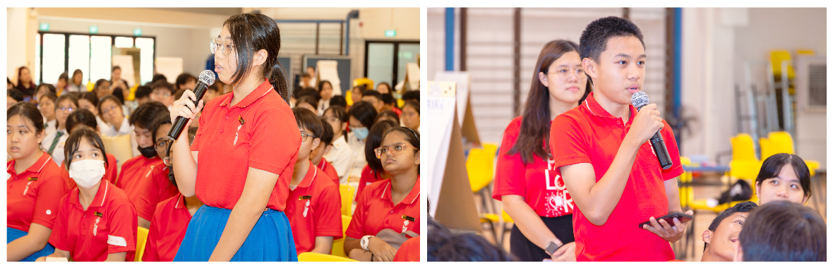 Two students in red uniforms each hold a microphone and speak before a large seated audience in a school hall.