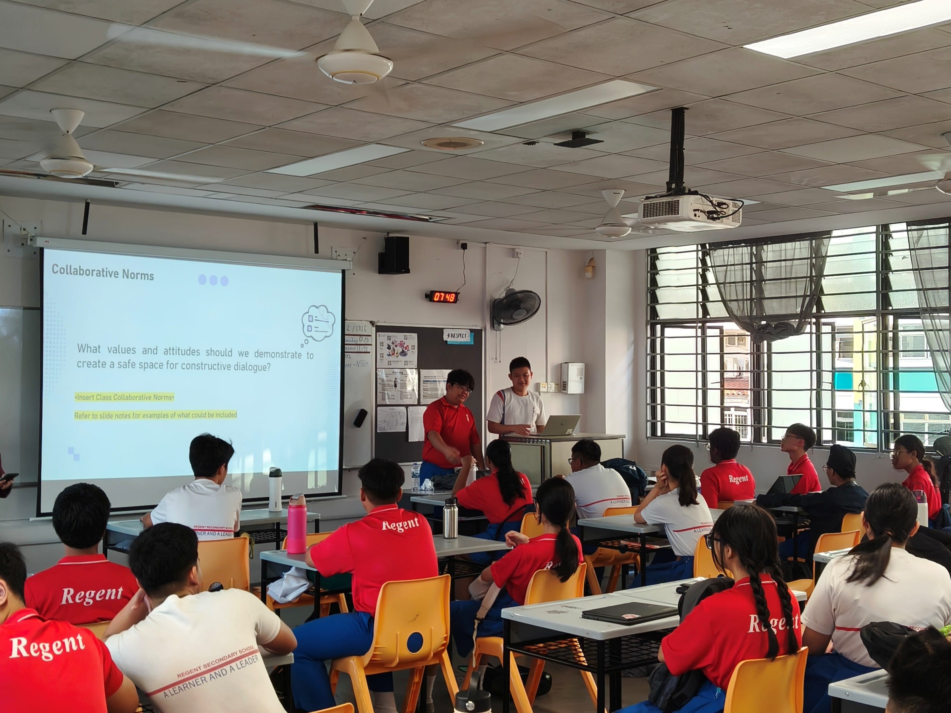 Students in red Regent uniforms sit at desks facing two presenters and a projected slide on Collaborative Norms.