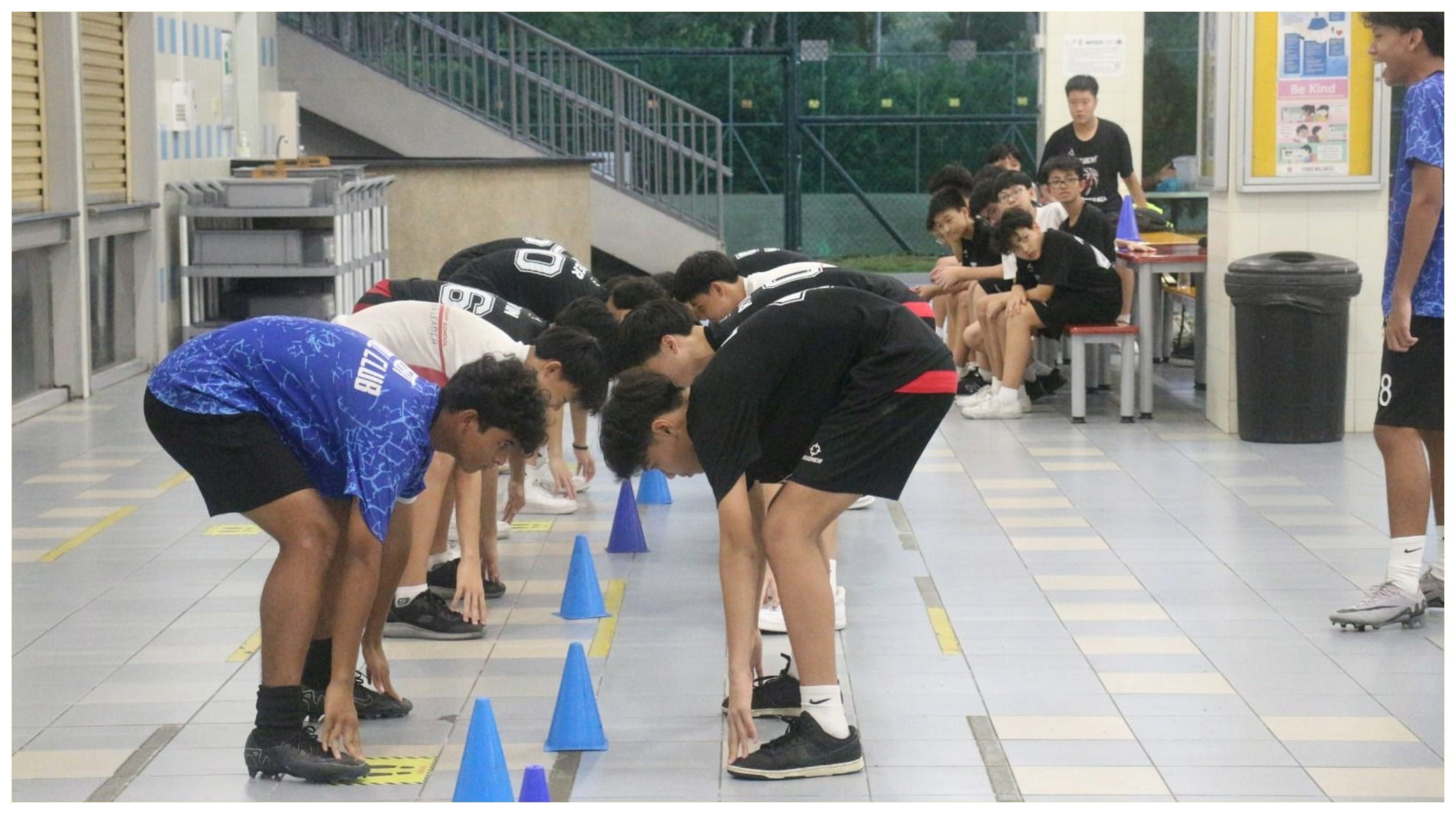 Several teenage boys bend down near blue cones on a tiled floor indoors, while others sit watching on benches.