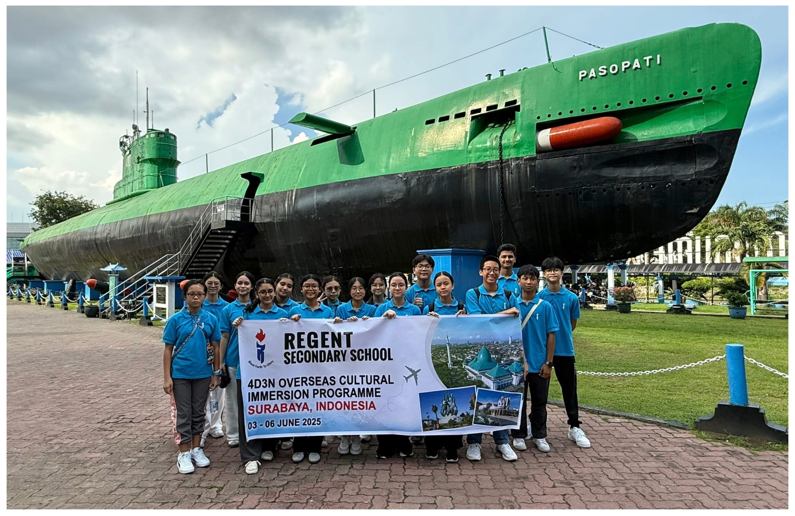 Students in blue shirts hold a Regent Secondary School banner in front of the green Pasopati submarine in Surabaya, Indonesia.