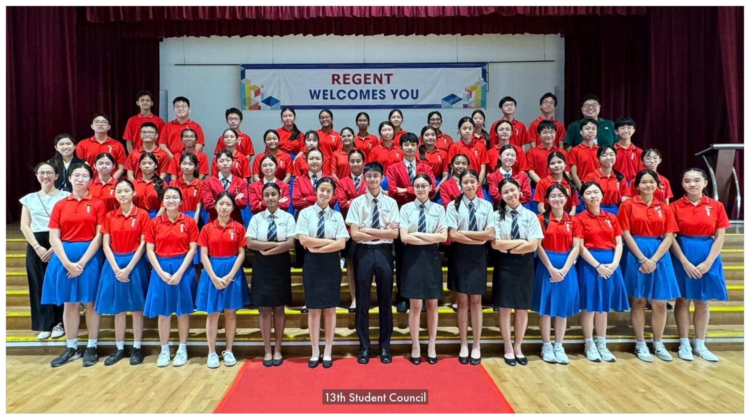 Large group of students in red and blue uniforms posing on a stage under a Regent Welcomes You banner.
