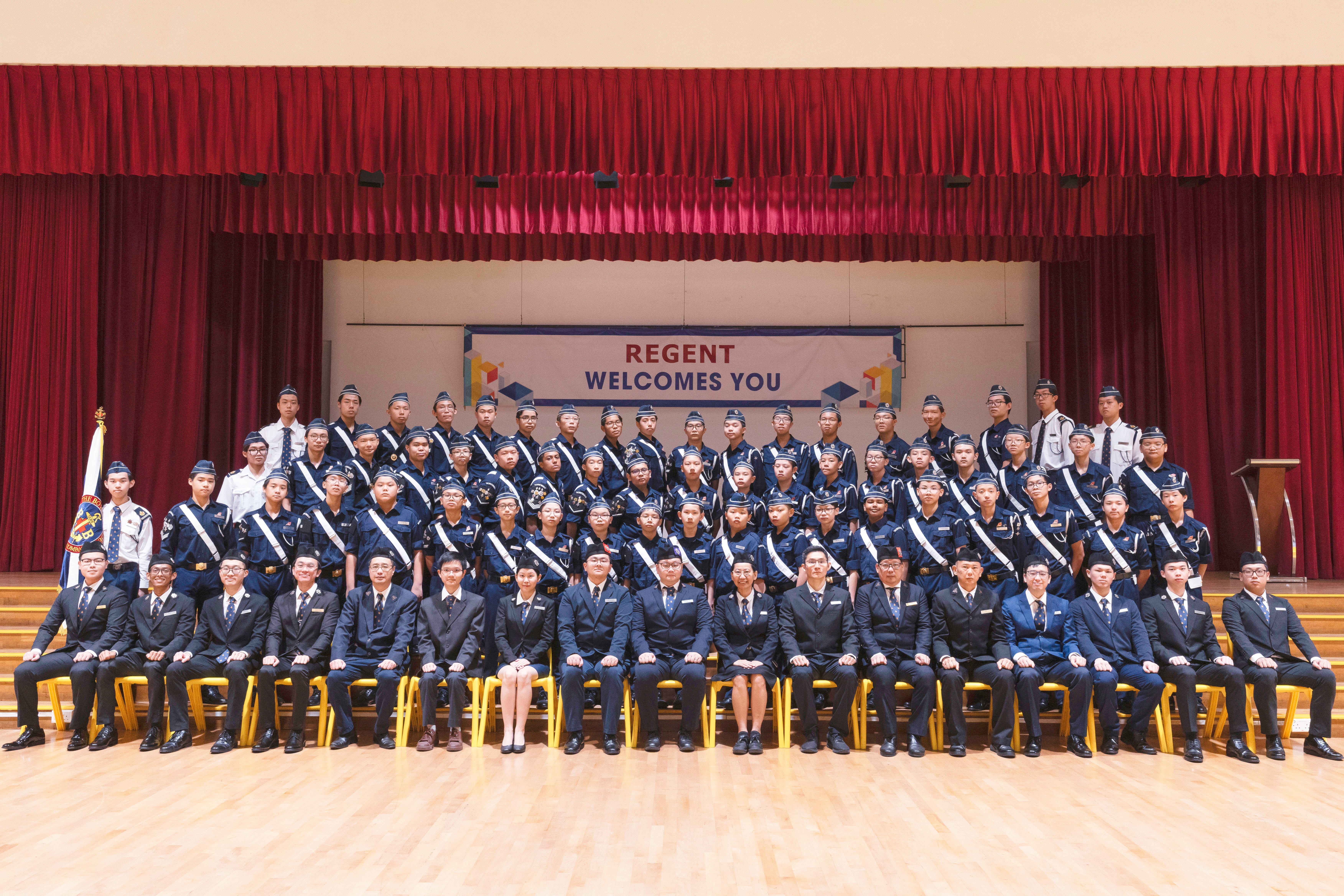 Large group in navy uniforms posed on stage under red curtains, banner reads REGENT WELCOMES YOU.
