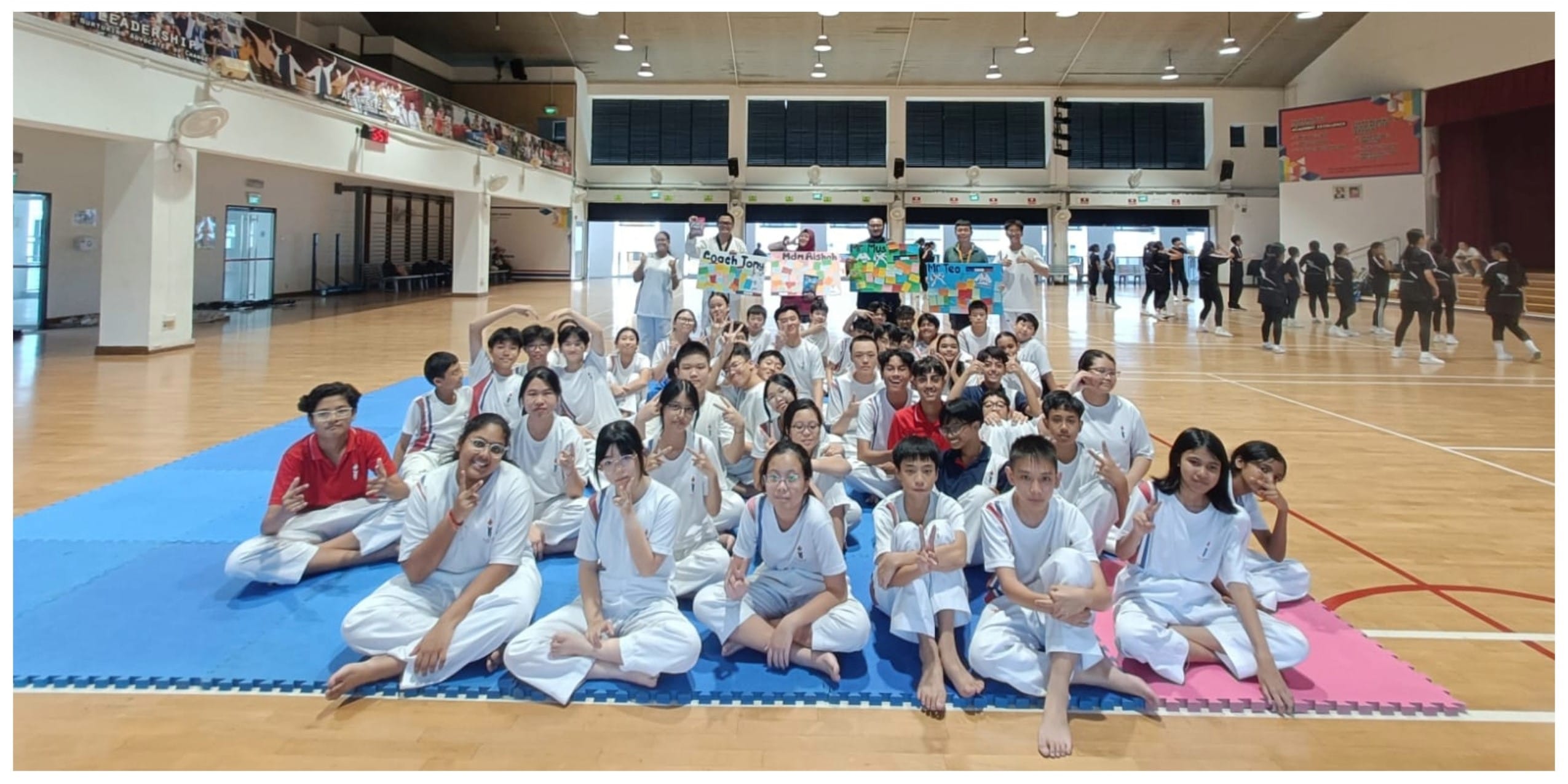 A large group of students in white uniforms pose on blue and pink mats in a school gymnasium.