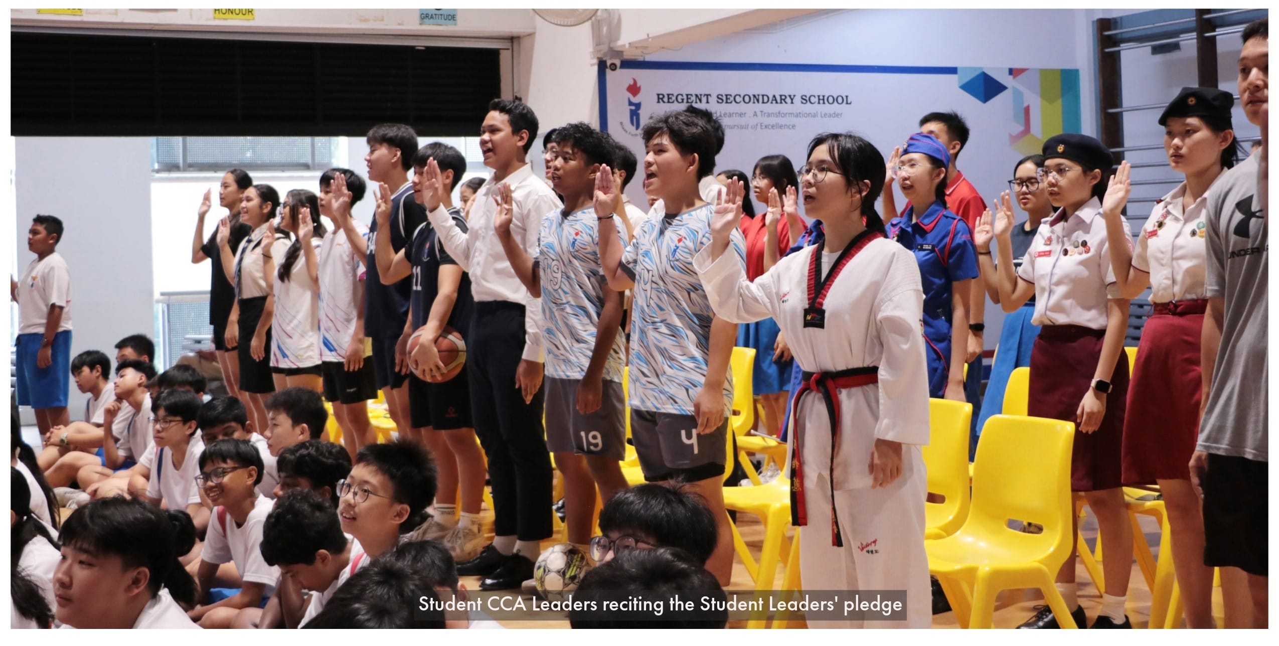 Students in various uniforms stand with raised hands reciting a pledge at Regent Secondary School.