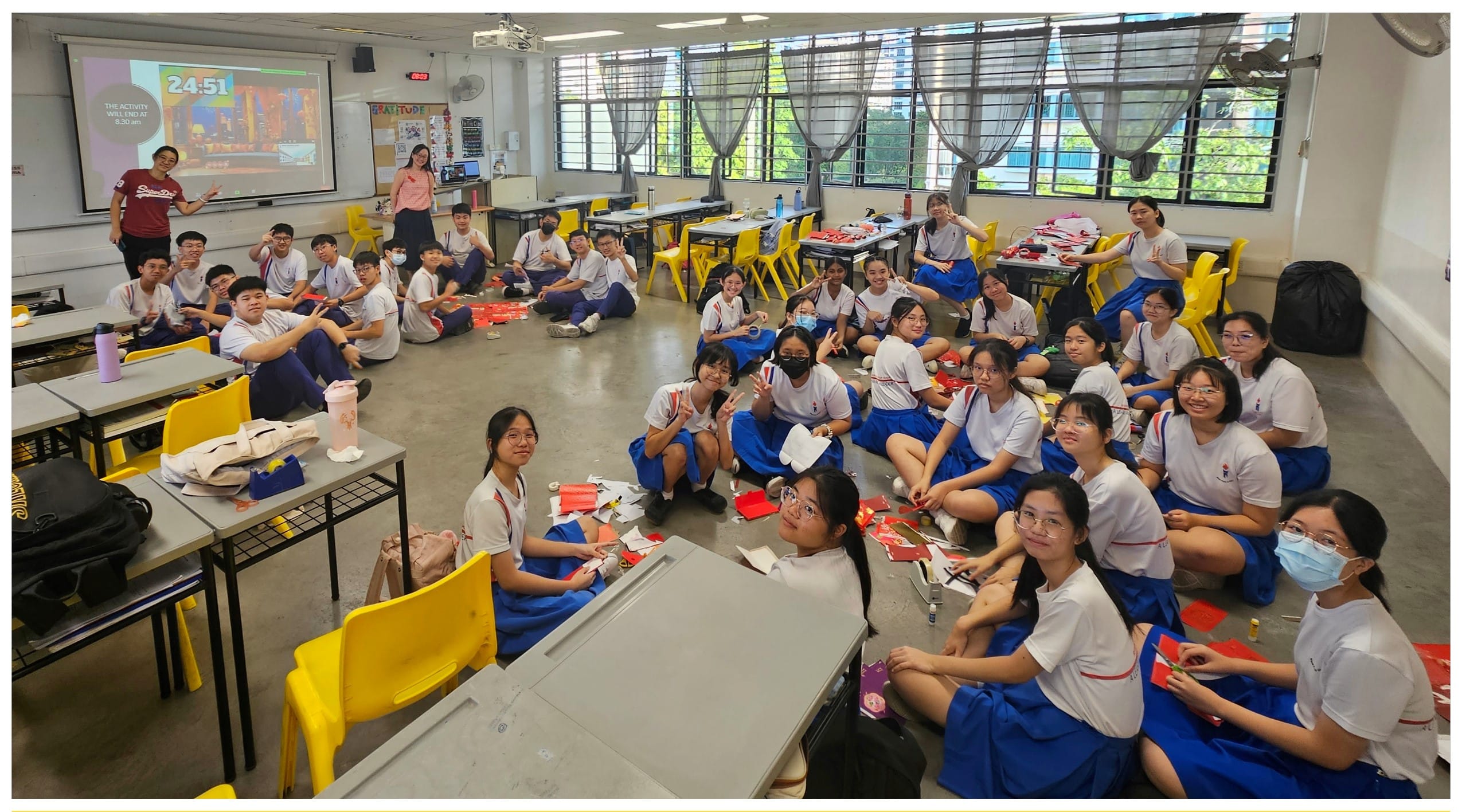 Students in white and blue uniforms sit on a classroom floor doing a craft activity with red paper envelopes.