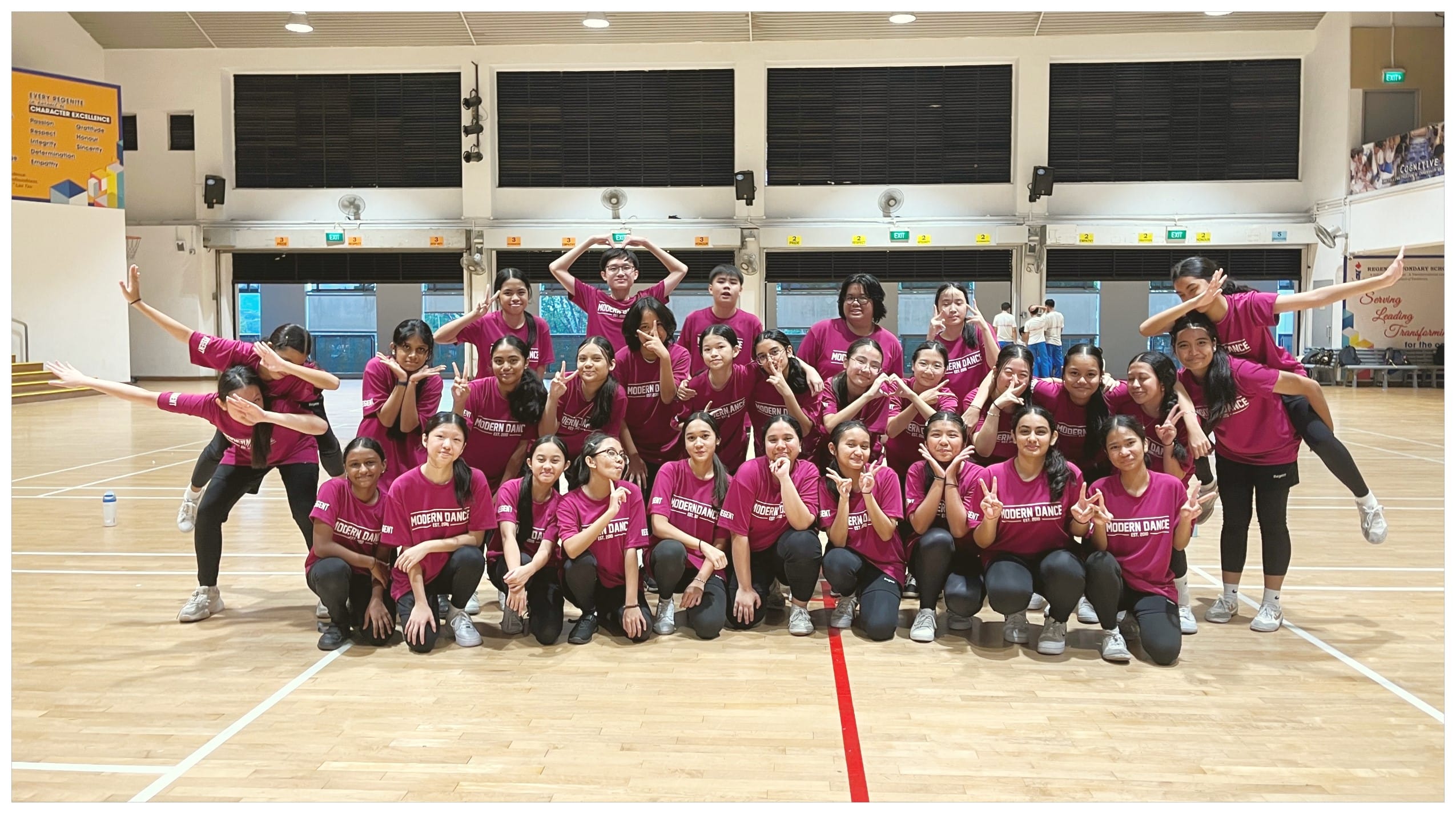 A large group of students in matching magenta Modern Dance shirts pose together in a school gymnasium.