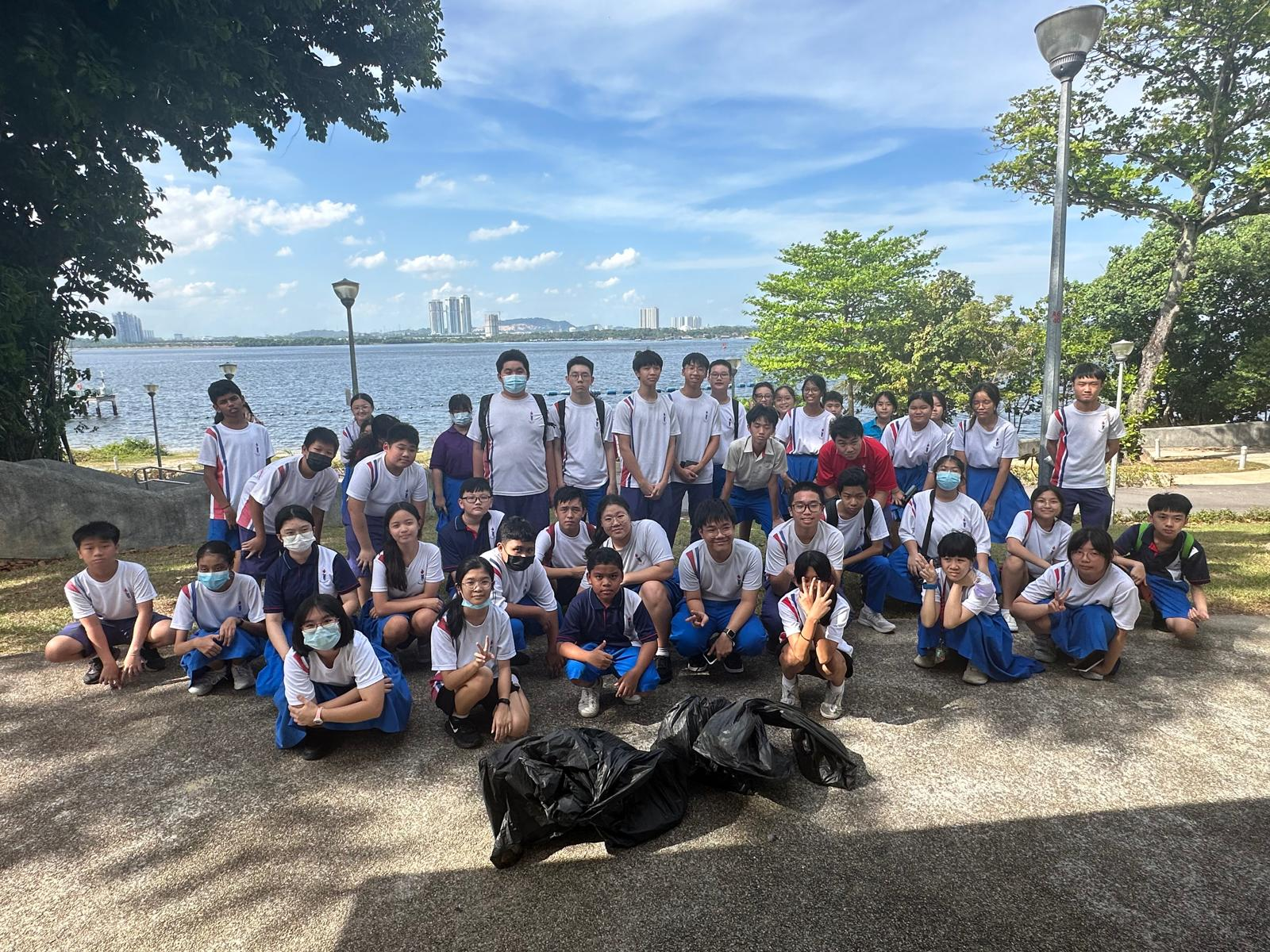 A large group of students in white and blue uniforms pose outdoors by a waterfront with filled black trash bags in front.