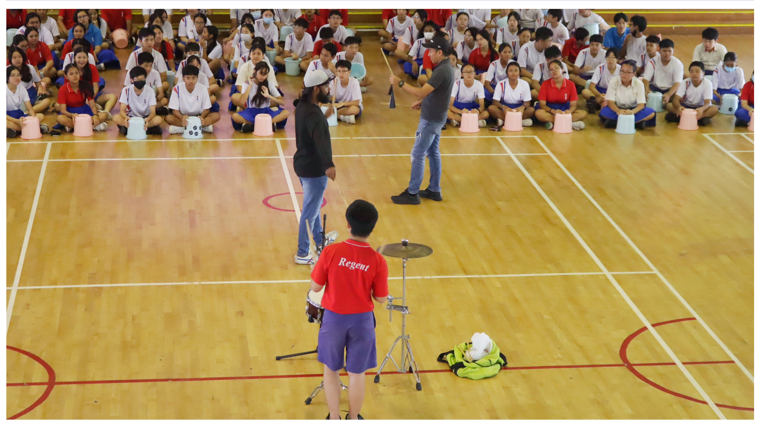 Students sit in a gym while two men speak at center; a student in red stands by a cymbal stand.