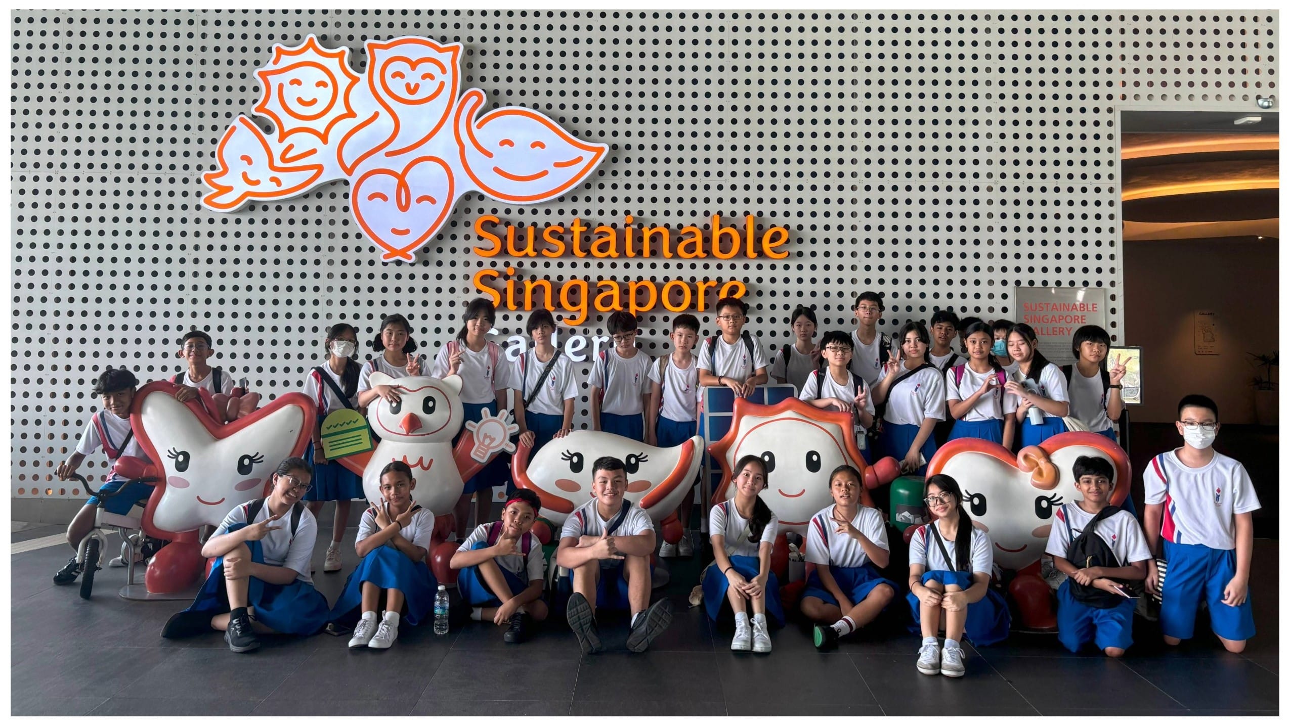 A group of students in school uniforms pose with cartoon mascots in front of the Sustainable Singapore Gallery sign.