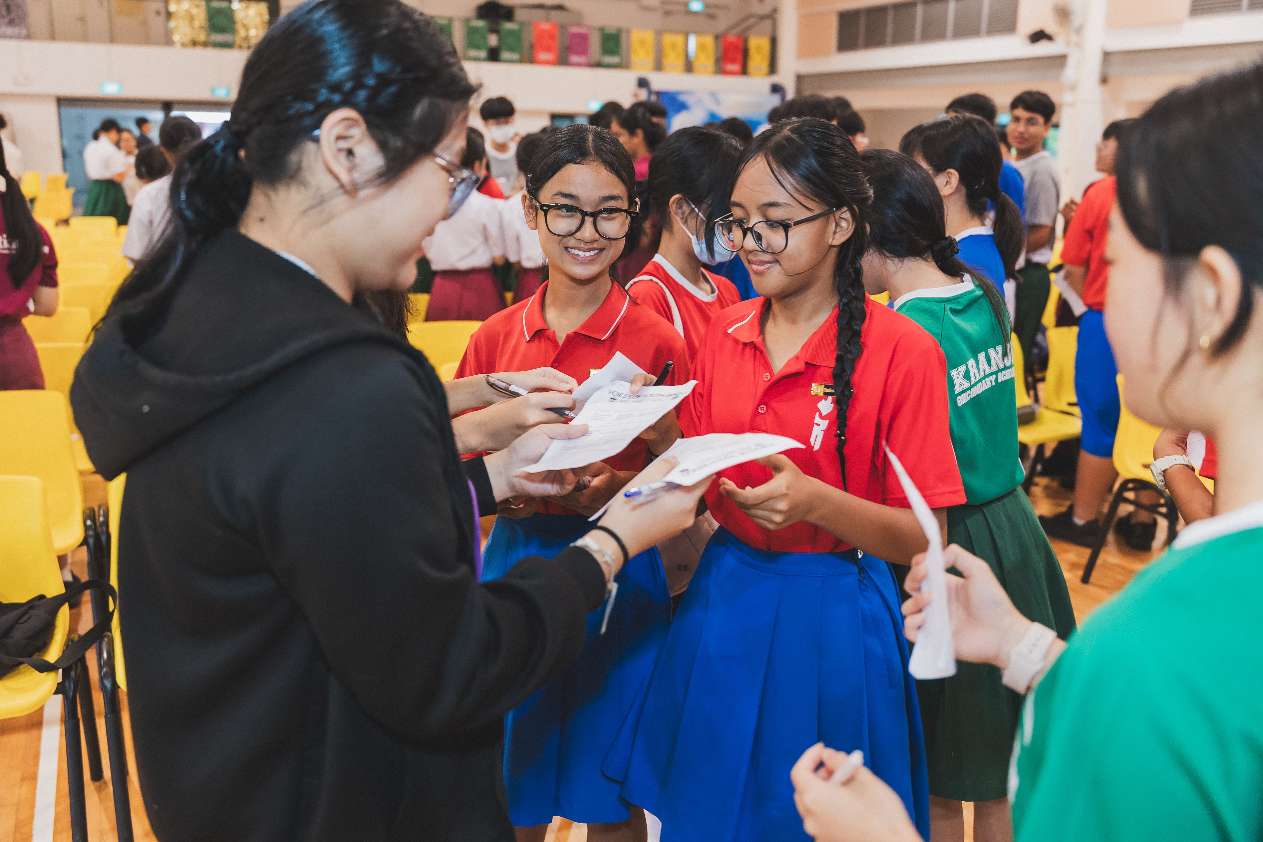 Students in colorful school uniforms gather indoors, smiling and exchanging papers during a school event.