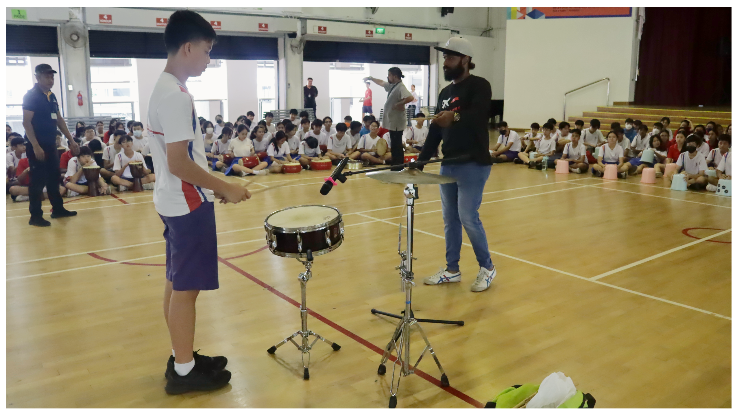 Student stands by a snare drum as an instructor demonstrates cymbal technique in a gym with seated classmates.
