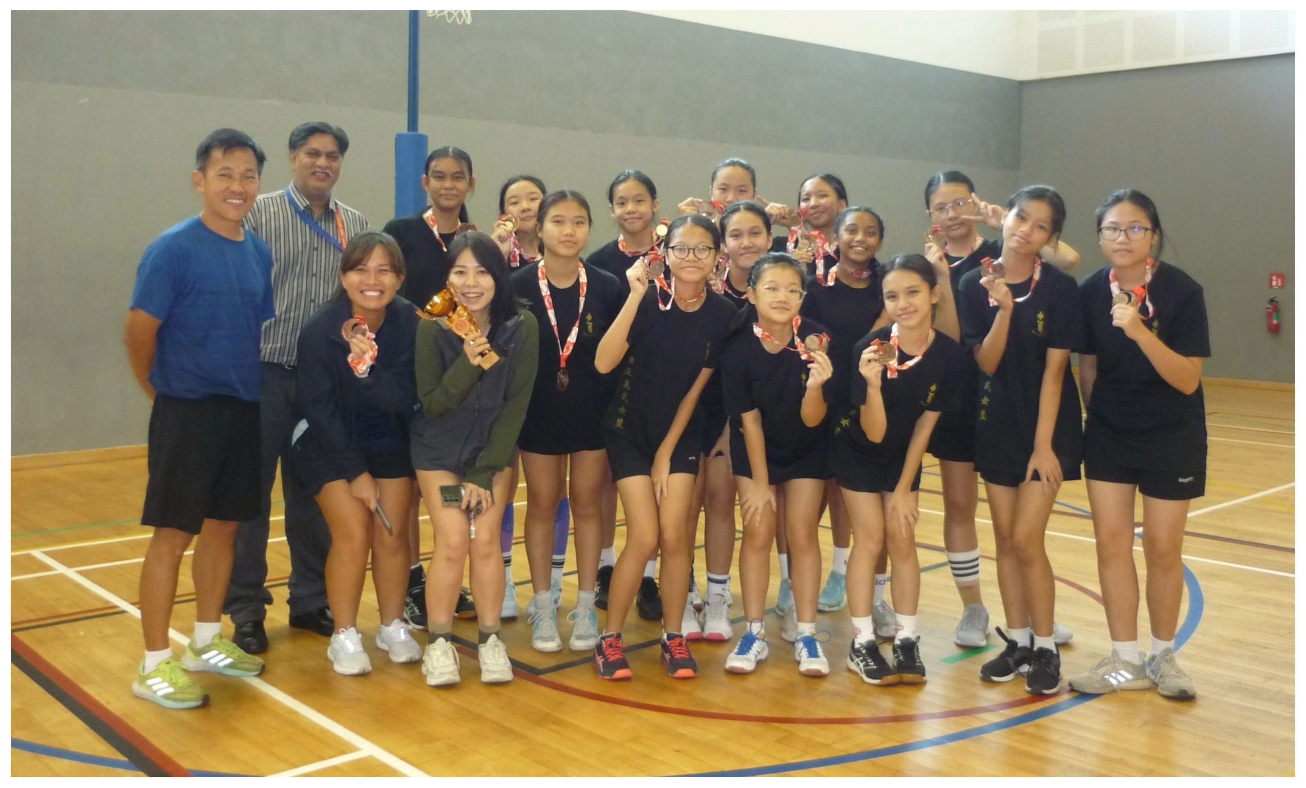 A group of young female athletes in black uniforms proudly display medals and a trophy inside an indoor sports hall.