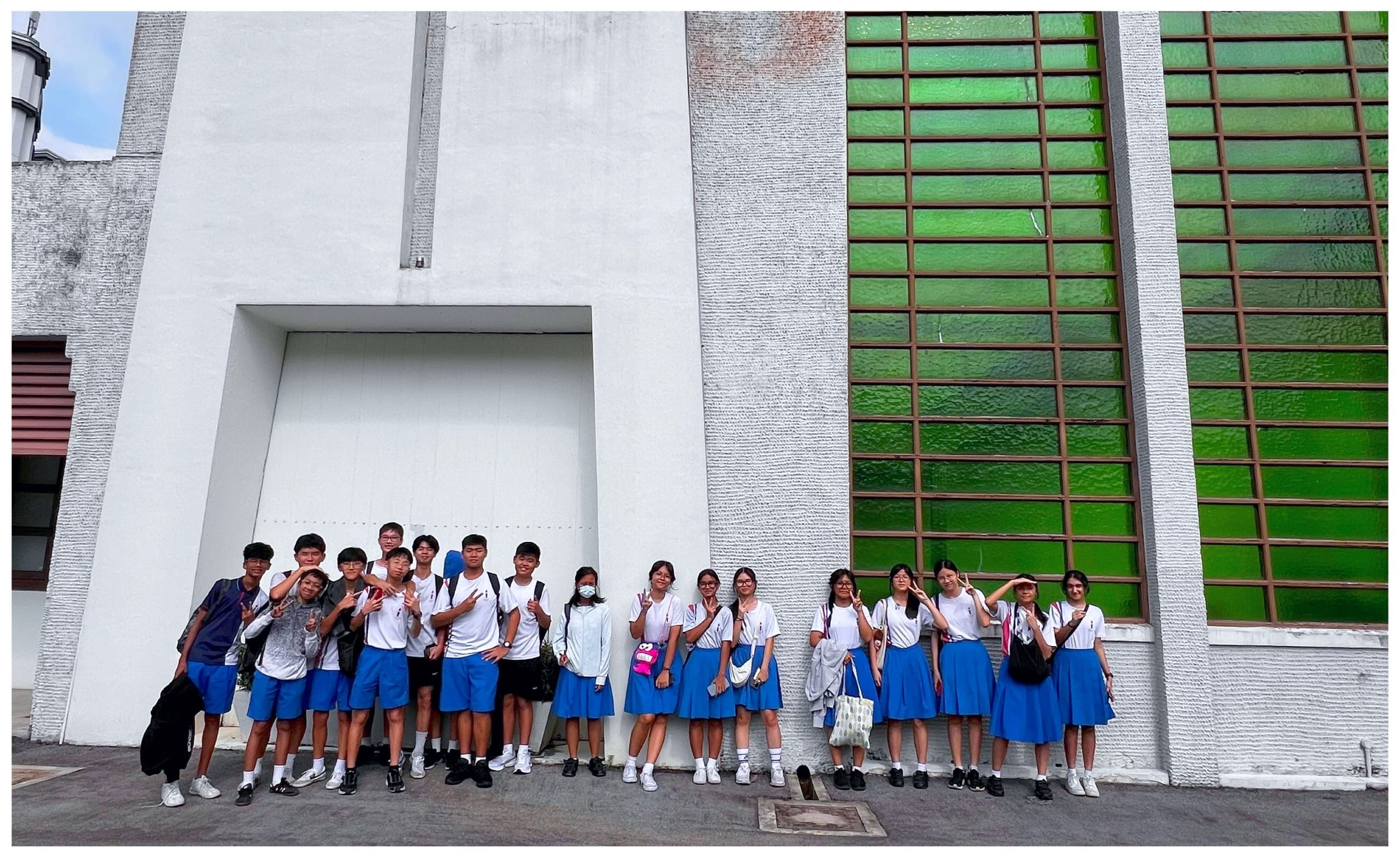 A group of students in white tops and blue uniforms pose outside a white building with large green glass windows.