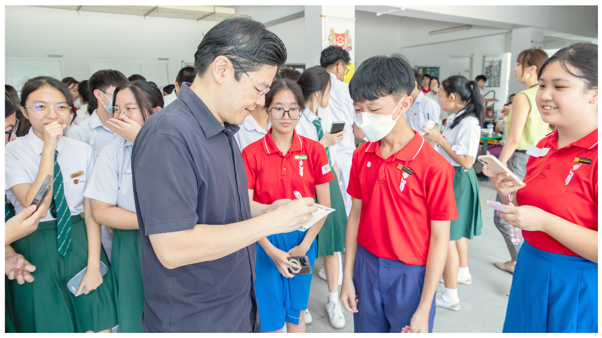 An adult man signs a card for school students in red and white uniforms gathered around him indoors.