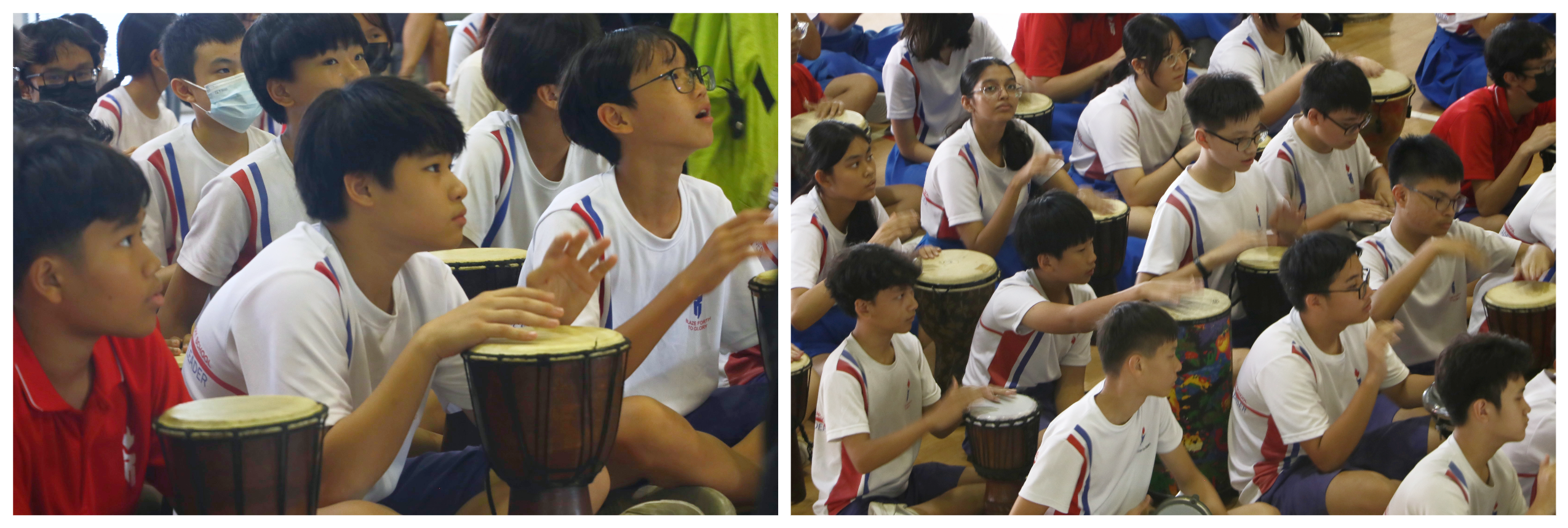 Students in white uniforms sit on the floor playing djembe drums during a group drumming session.