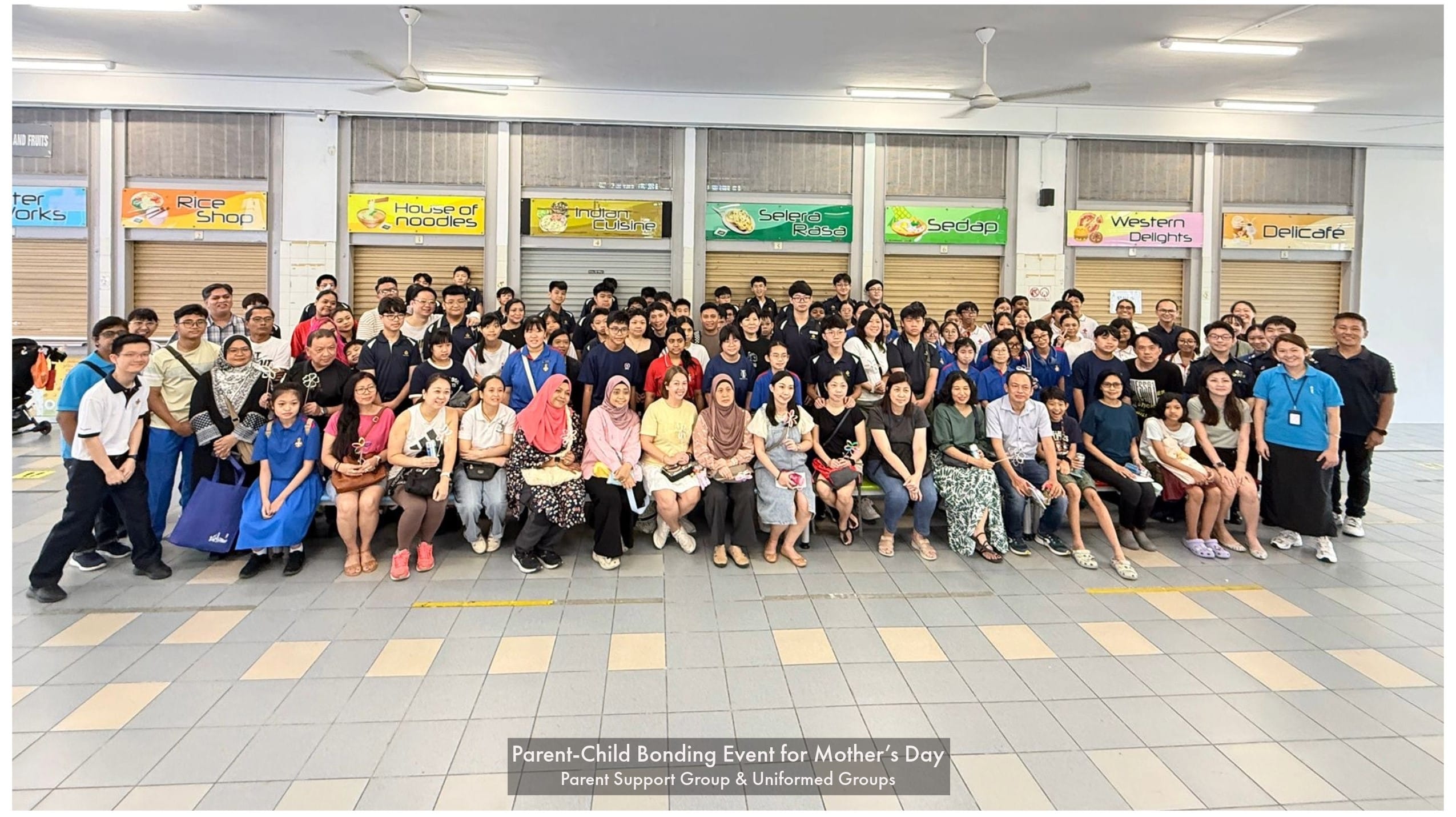 Large group of adults and students posing indoors at a school canteen for a Mother's Day bonding event.