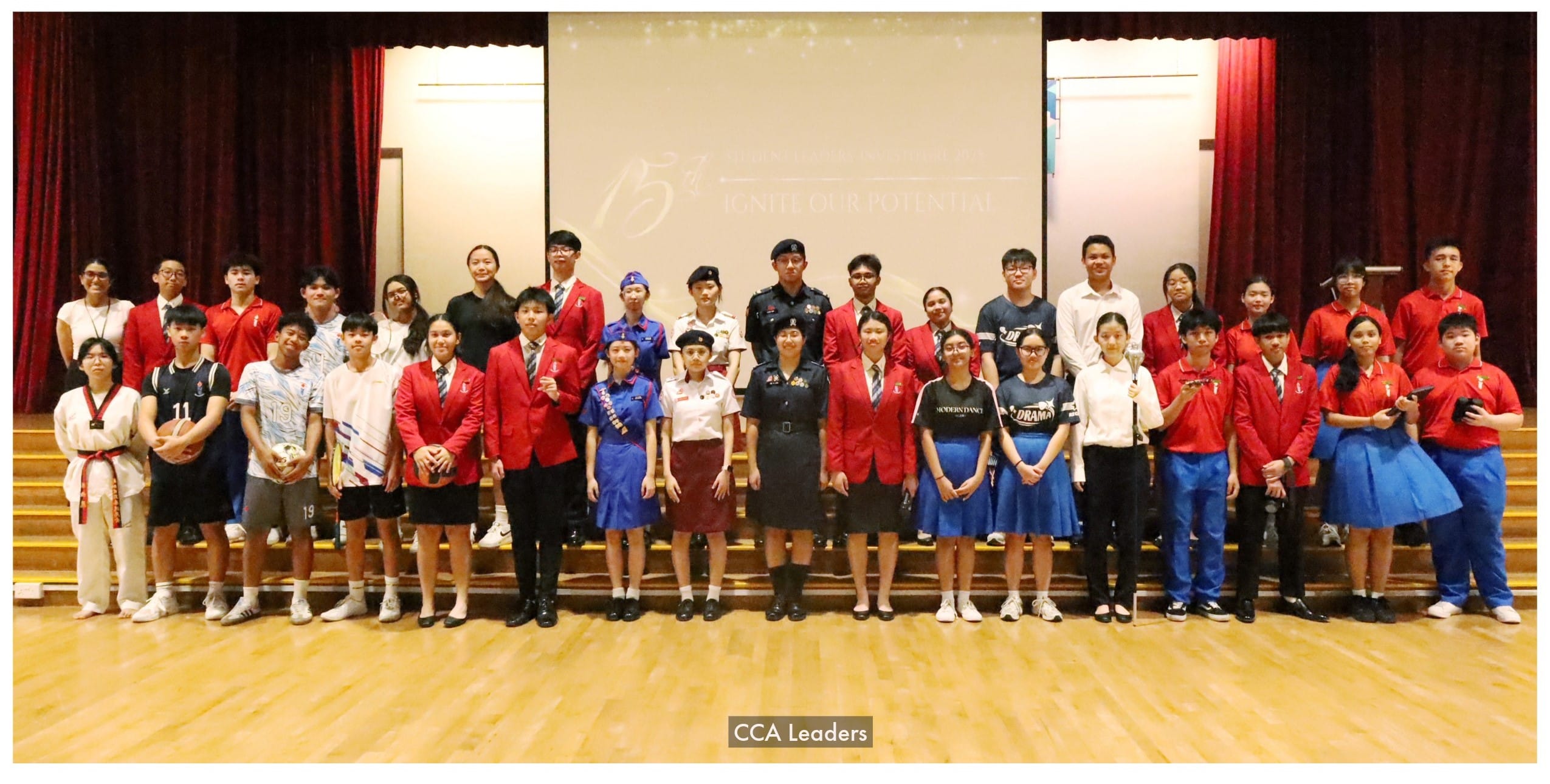 Group of students in various uniforms and activity attire posing on a school hall stage labeled CCA Leaders.