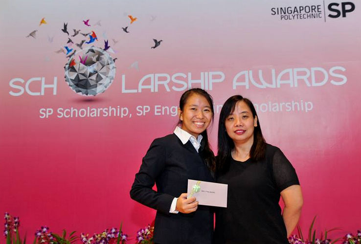 Two women pose smiling at a Singapore Polytechnic Scholarship Awards backdrop, one holding an envelope.