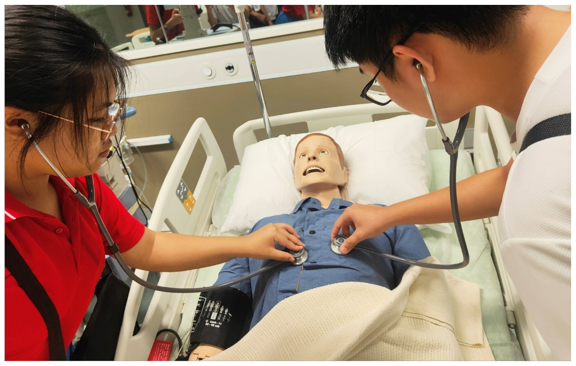 Two students use stethoscopes on a medical training mannequin lying in a hospital bed.