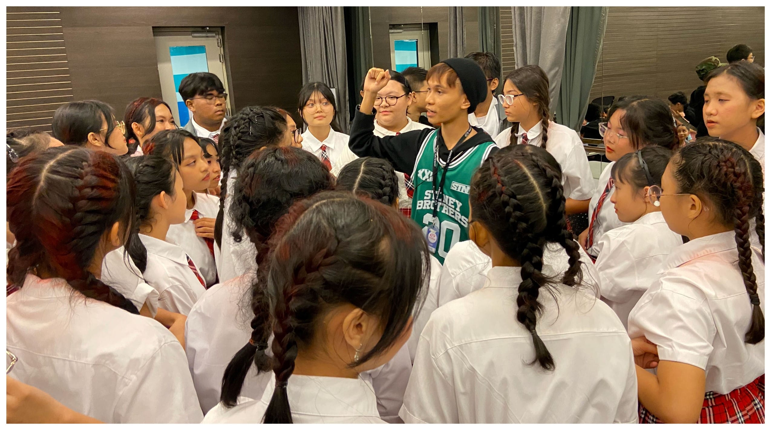 A person in a green jersey and black beanie speaks to a large group of students in white uniforms indoors.