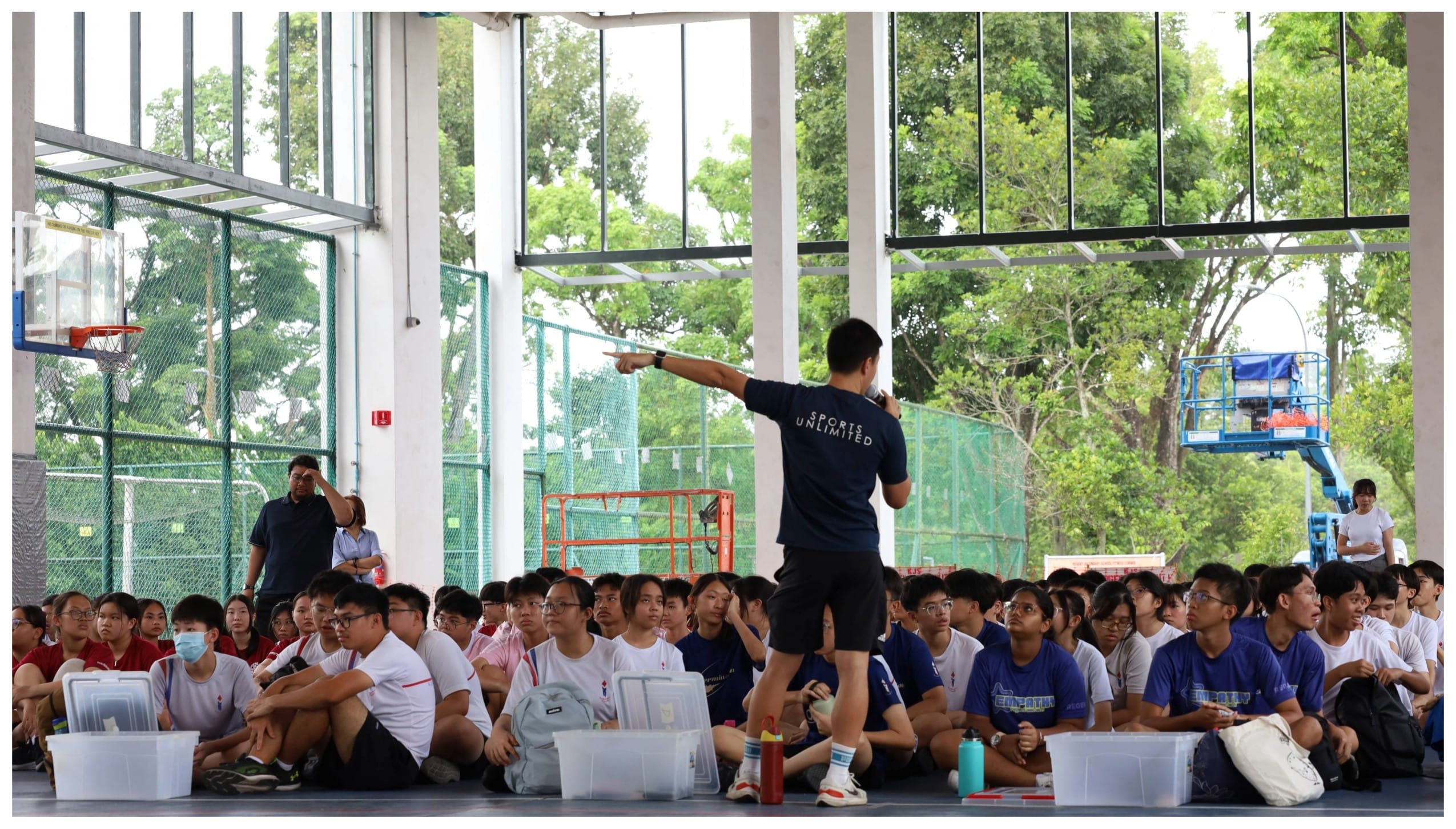A person in a Sports Unlimited shirt points while addressing a large group of students seated on a gym floor.
