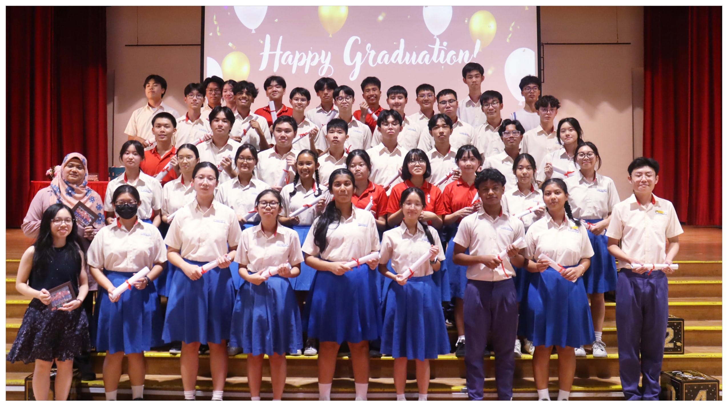 Large group of students in school uniforms holding diploma scrolls posing in front of a Happy Graduation banner.