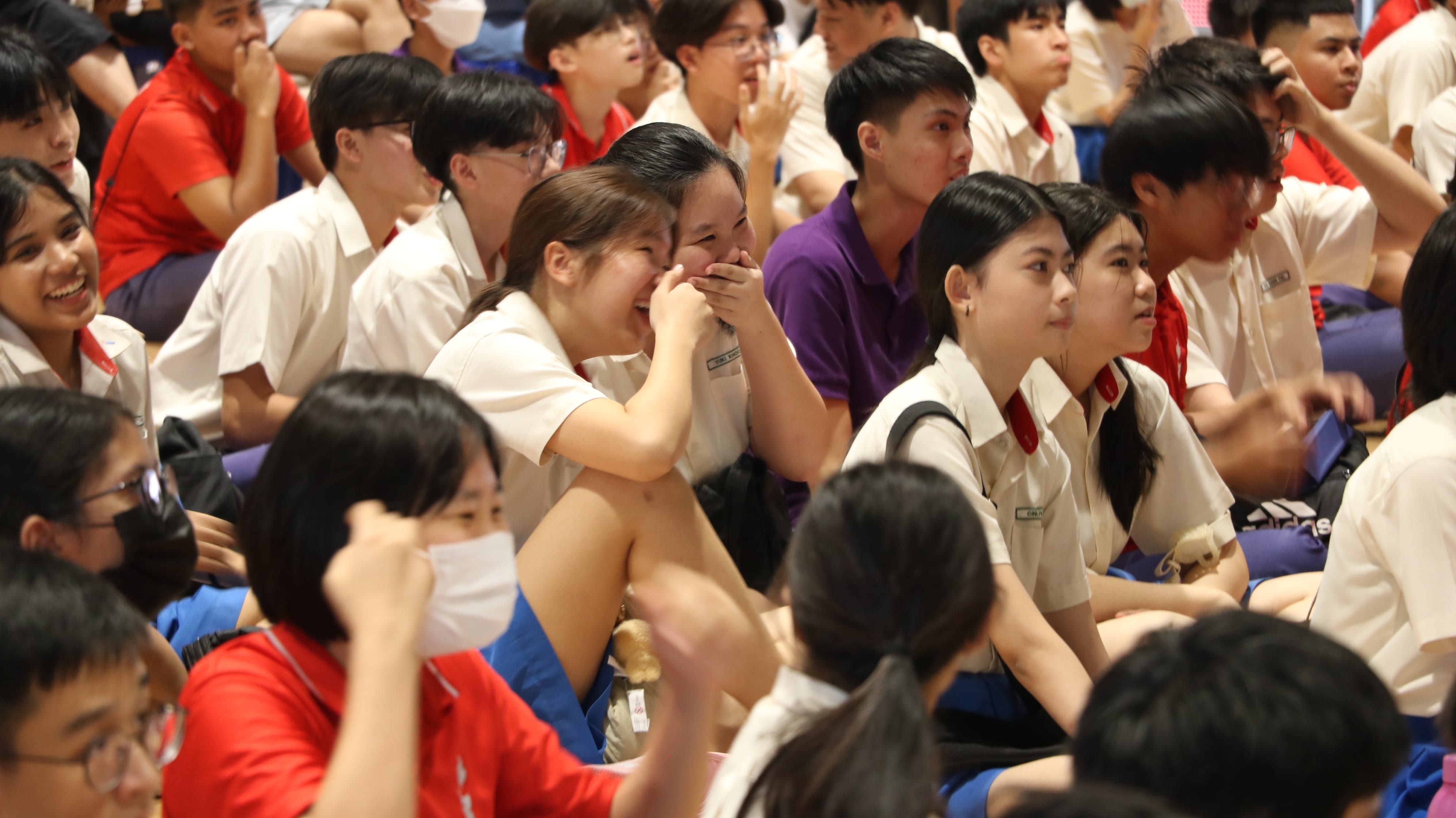 A large group of school students in cream uniforms sit together, with some laughing and smiling during an assembly.