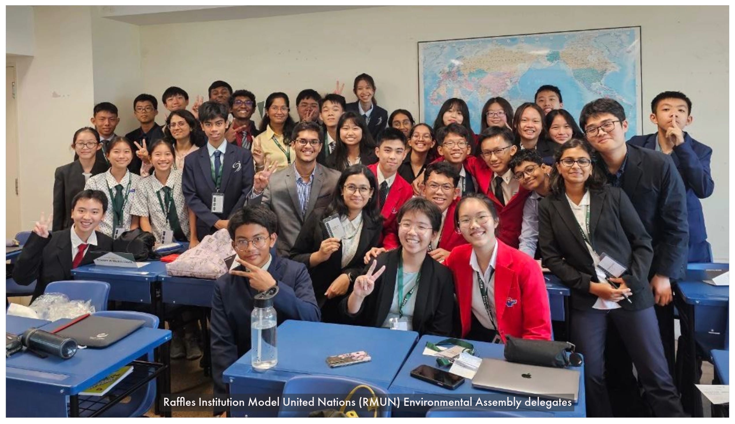 A large group of students in formal attire pose smiling in a classroom with a world map on the wall.