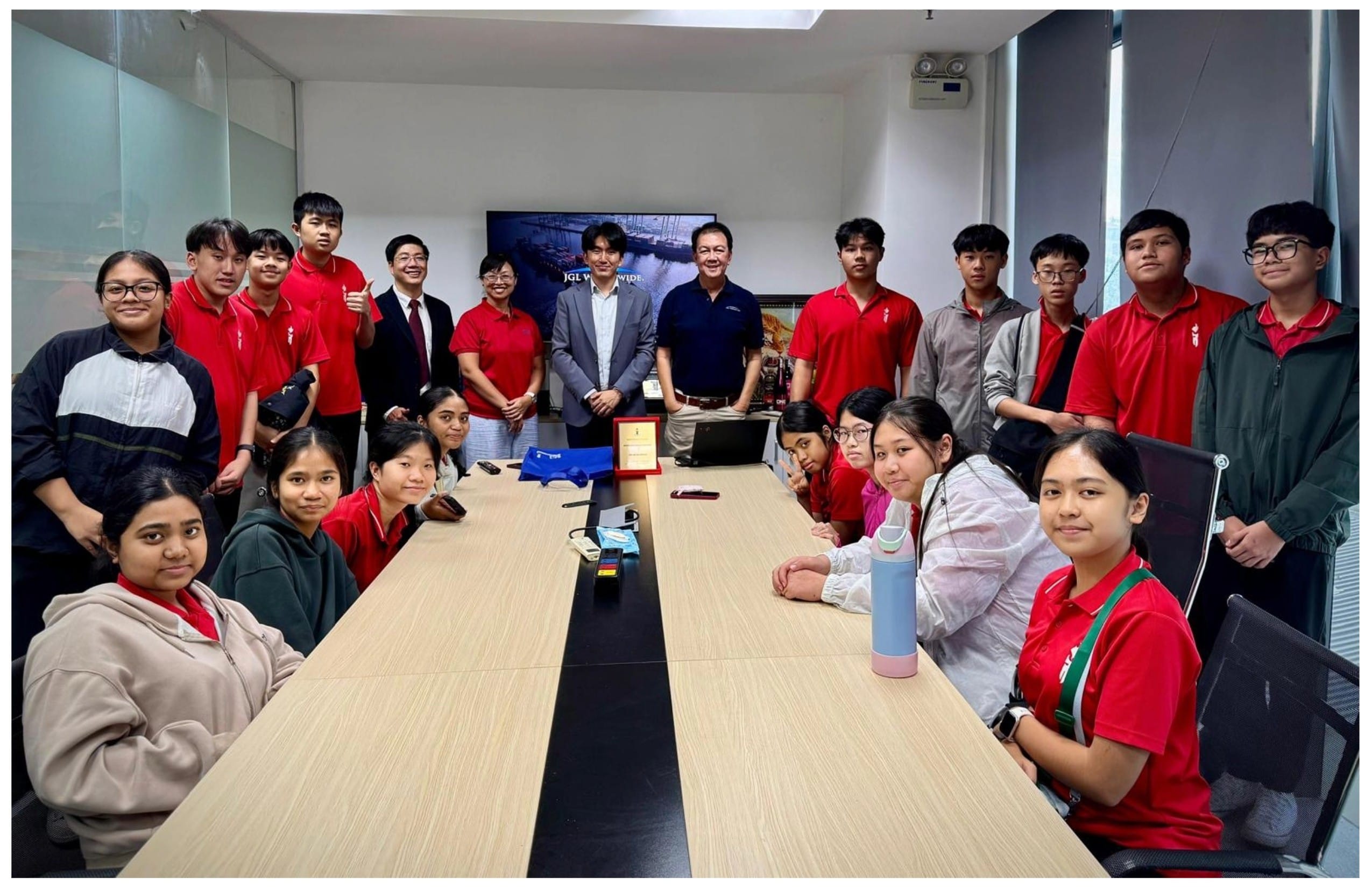 A group of students in red uniforms and two adults in suits pose together around a conference table in an office.