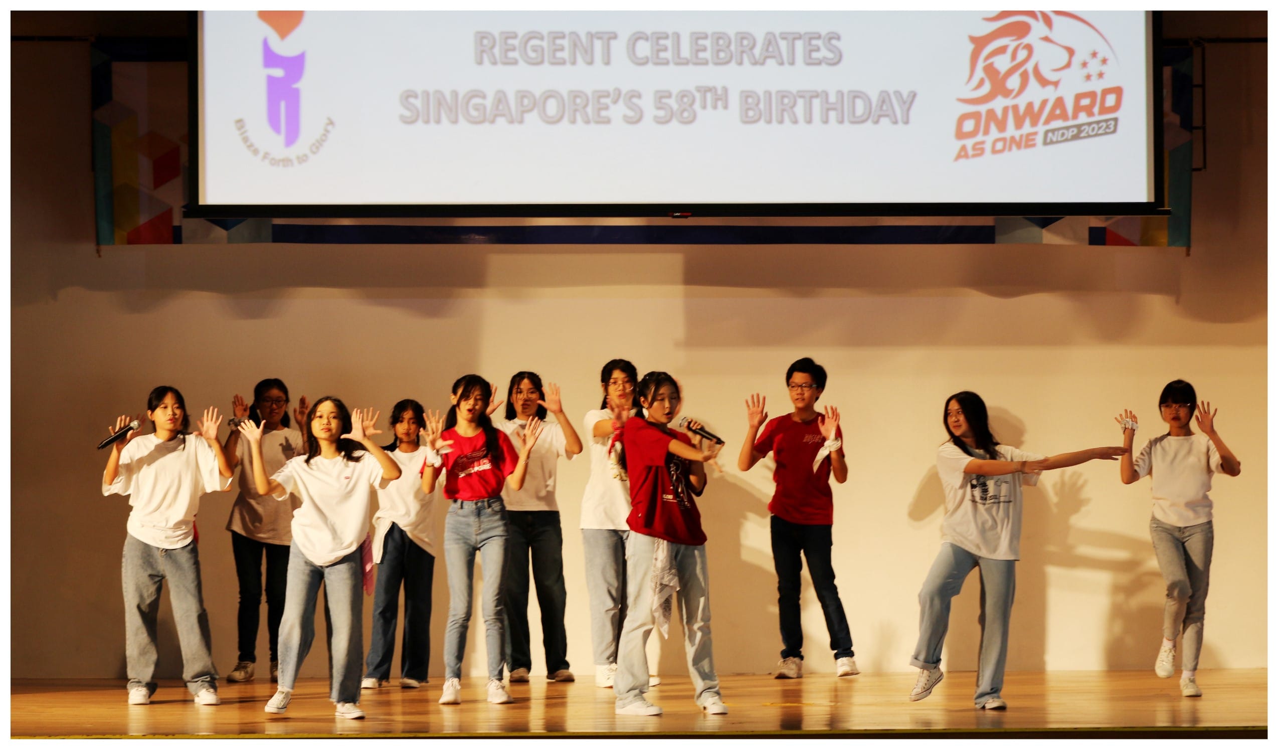 Students in white and red shirts perform on stage under a screen reading Regent Celebrates Singapore's 58th Birthday.