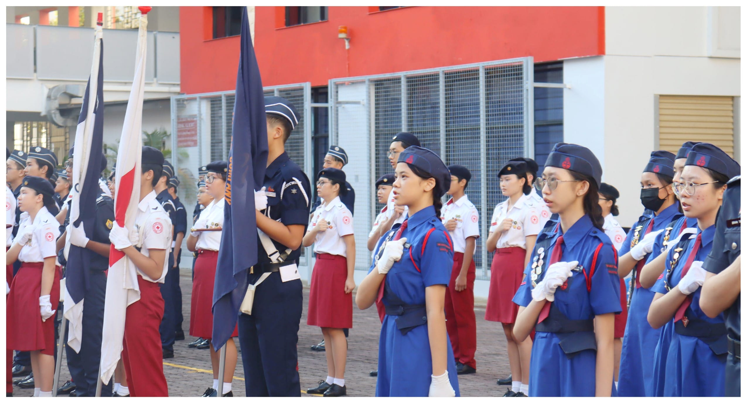 Students in navy and white uniforms stand at attention during a outdoor parade ceremony, holding flags near a red building.