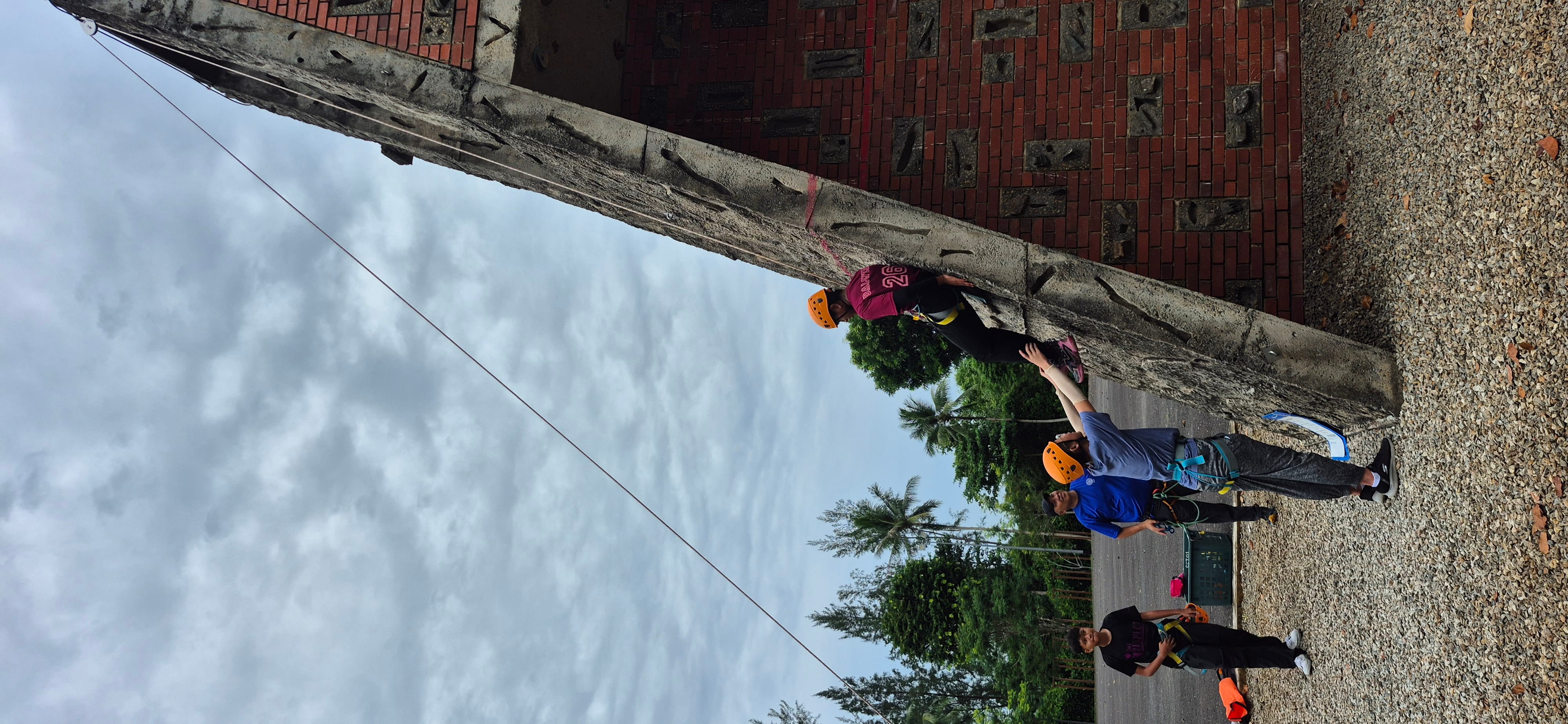 A person in an orange helmet climbs an outdoor rock wall while two people below assist, with trees and cloudy sky behind.