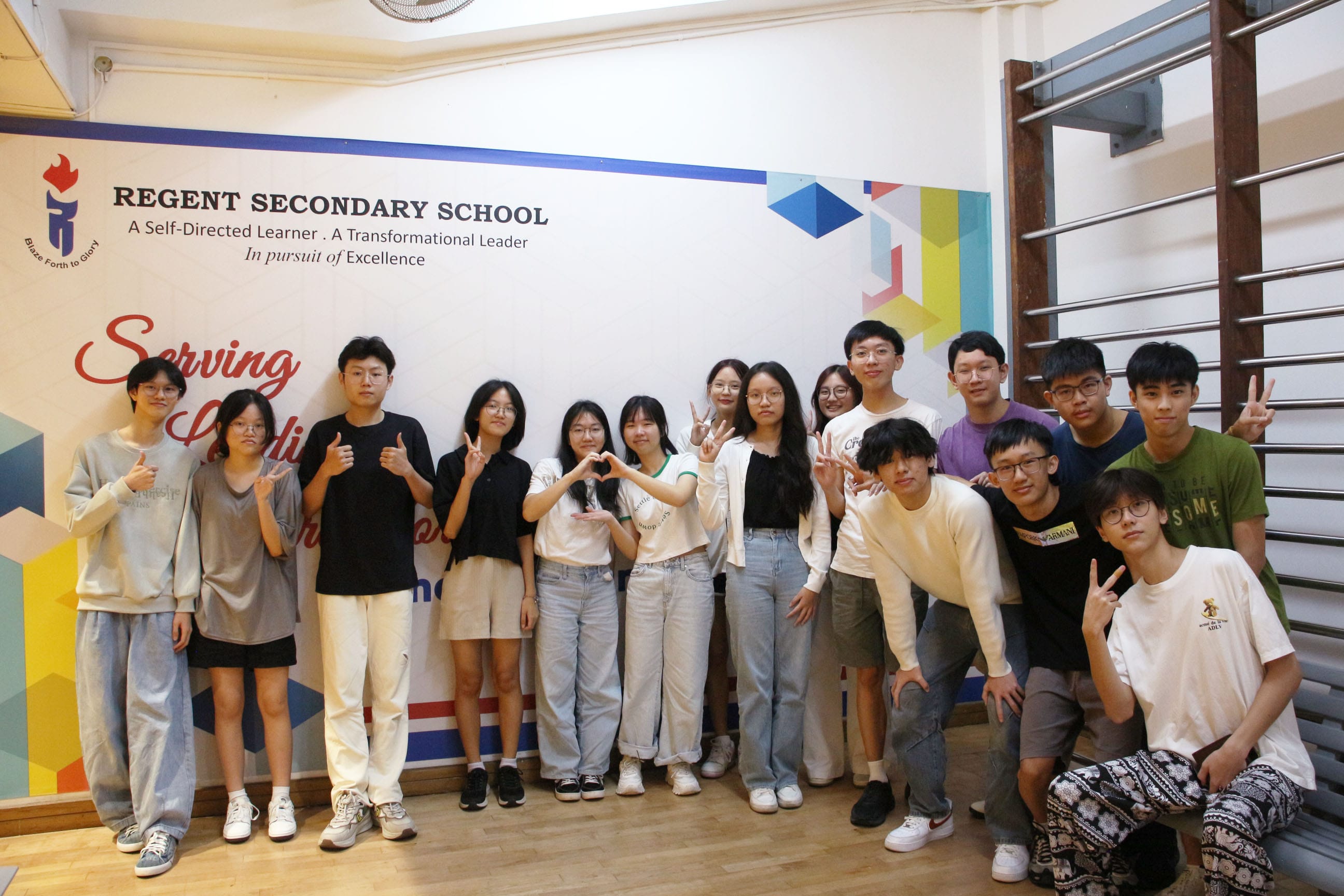 A group of teenagers posing in front of a Regent Secondary School banner, making peace signs and heart gestures.